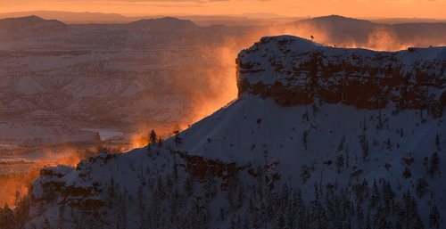 A shadow covered rock formation covered in snow with frost blowing in the wind illuminated orange by the sun.