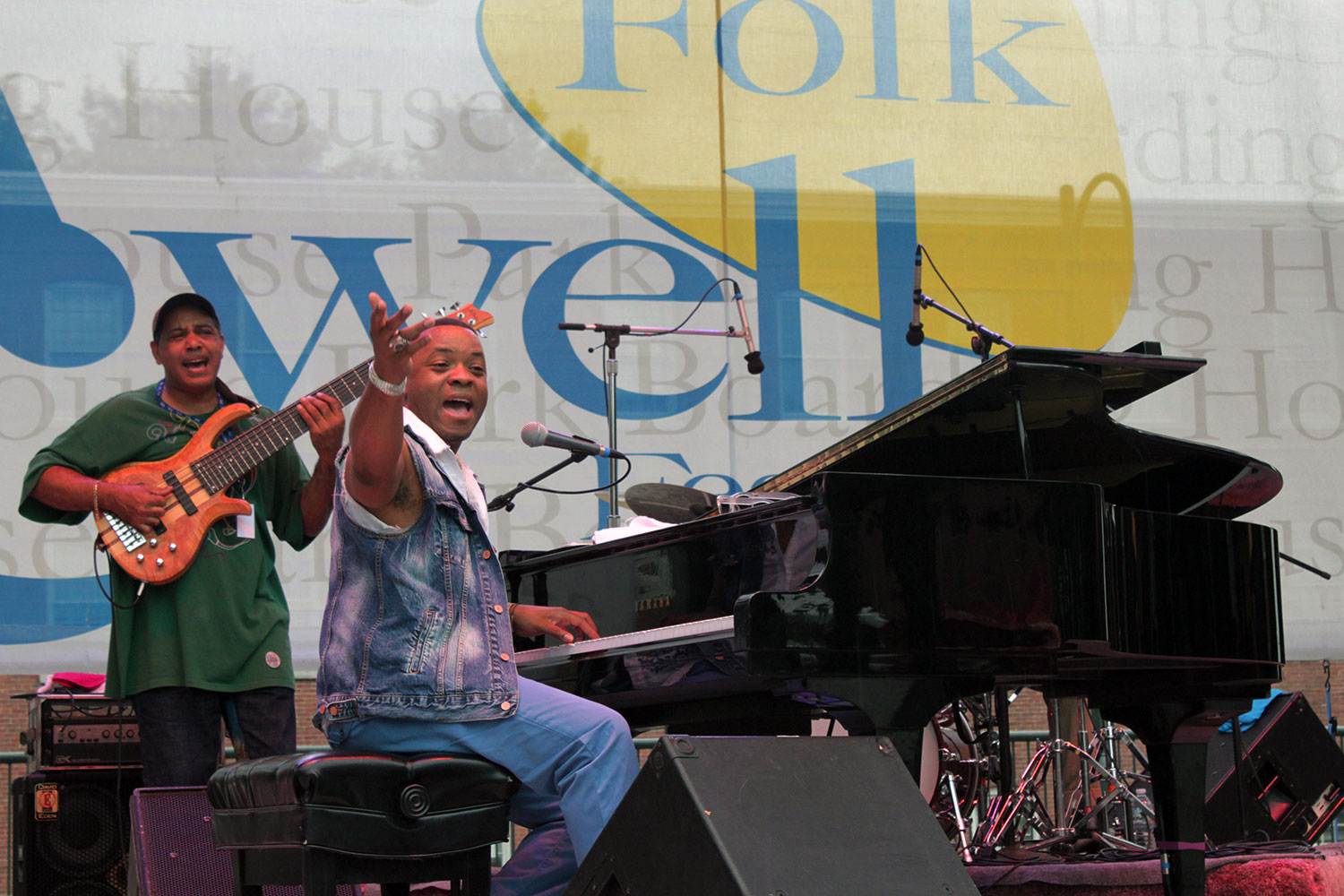 On stage, Davell ushers his hand towards the crowd while seated playing the piano. He wears jeans, and a sleeveless denim jacket. Further back on the stage, Severan is playing bass guitar.