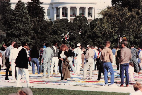A collection of people stands on multi-colored cloth laid on the grass. They are looking down at the cloth. Trees and the south lawn of the White House are visible in the background
