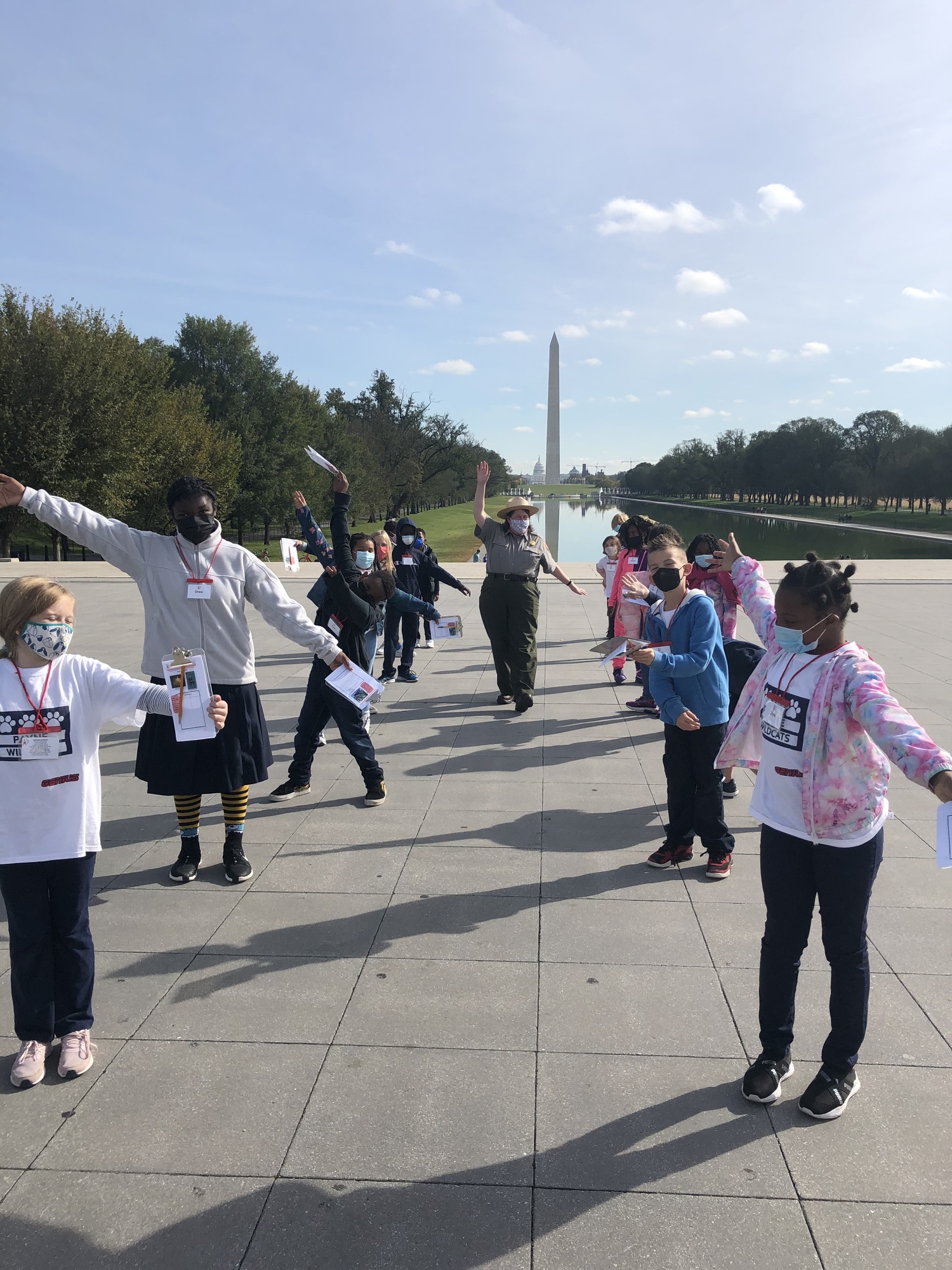Park ranger in uniform raises right arm and left arm down as students copy her movement. Washington Monument and reflecting pool of water in the background.