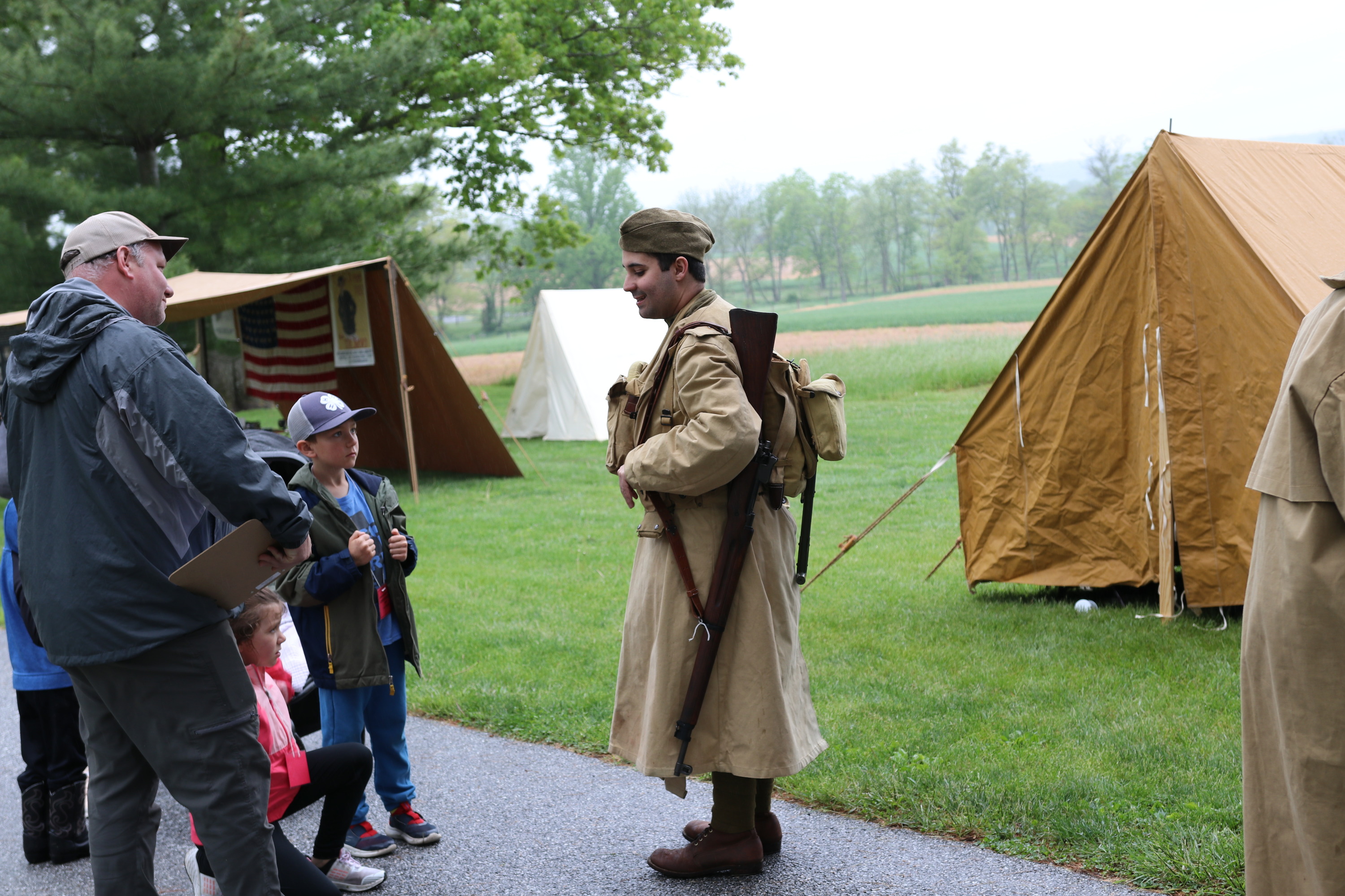 A living historian is dressed in period clothing for the WWI era and chats with a young visitor and an adult. There are two tents set up in the background of the photo. 
