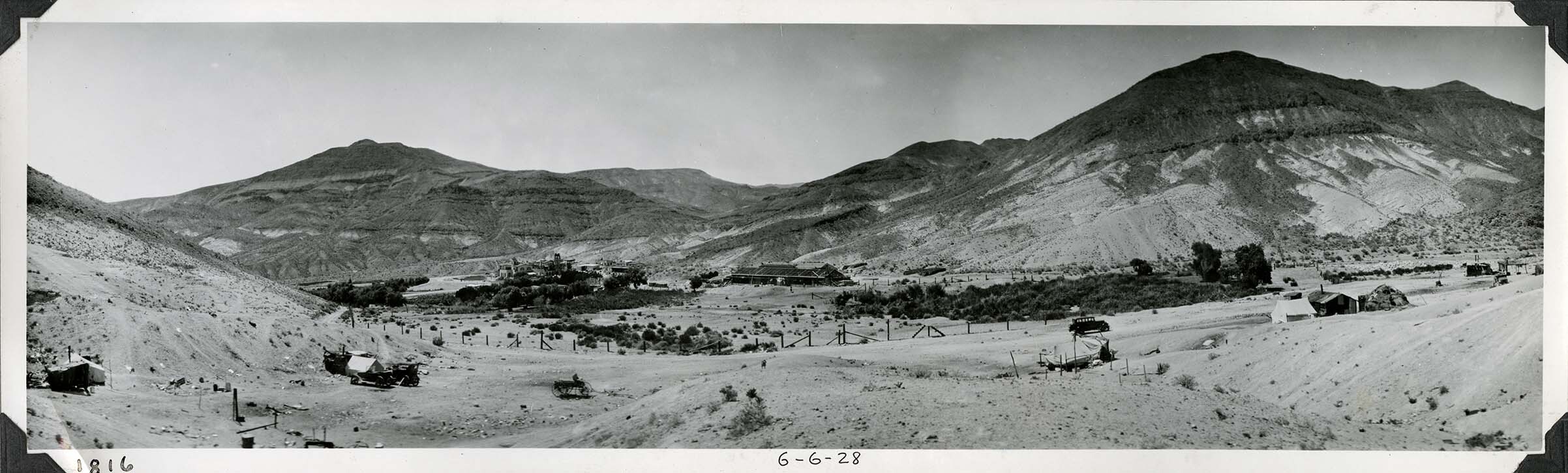 This is an historic black and white panoramic photograph from the Scotty's Castle Historic Photograph Collection, Death Valley National Park of a stark desert landscape with building and planted vegetation complex. Tent structures scattered across foreground. Dirt road with parked vehicle goes off to right. Fences pastures and vegetation in middle ground. Large building under construction in middle of image. Rest of Upper Grapevine Ranch structures to the left. Desert hills in background. Inscriptions in black ink along lower border.