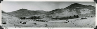 This is an historic black and white panoramic photograph from the Scotty's Castle Historic Photograph Collection, Death Valley National Park of a stark desert landscape with building and planted vegetation complex. Tent structures scattered across foreground. Dirt road with parked vehicle goes off to right. Fences pastures and vegetation in middle ground. Large building under construction in middle of image. Rest of Upper Grapevine Ranch structures to the left. Desert hills in background. Inscriptions in black ink along lower border.