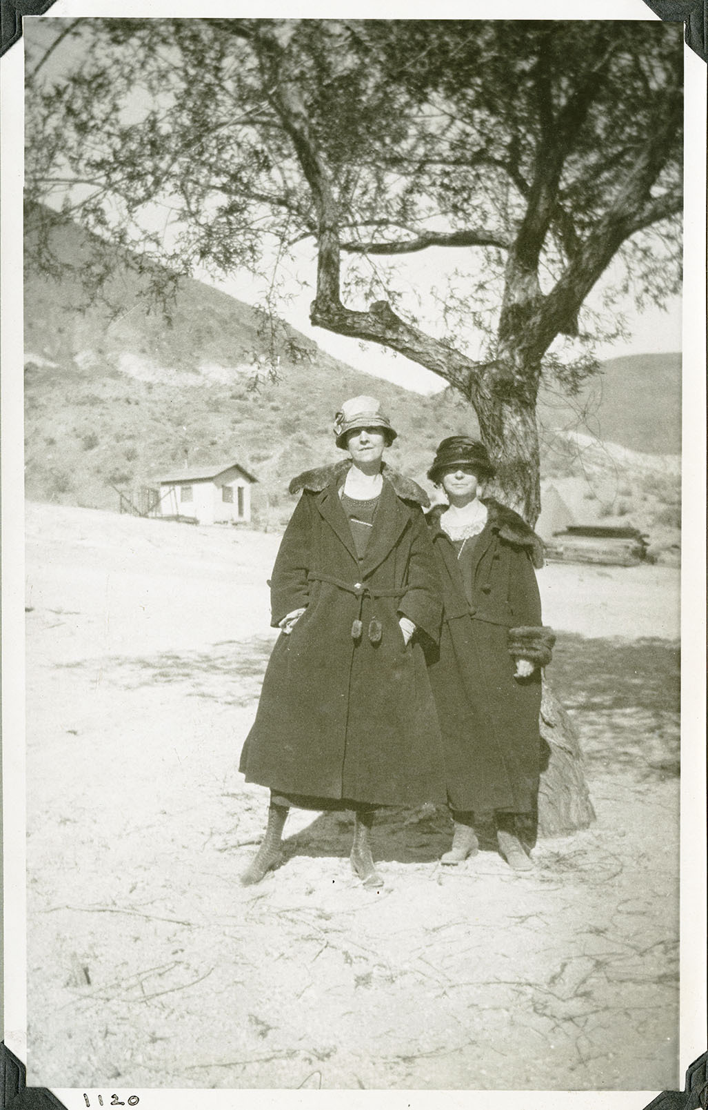 This is an historic black and white photograph from the Scotty's Castle Historic Photograph Collection, Death Valley National Park of two women posing for camera by a tree. Both dressed in 1920s style. Both have cloche hats, dresses, heavy fur-trimmed winter coats, high top laced shoes with heal. Simple stucco building behind. Number in black ink in lower left corner.