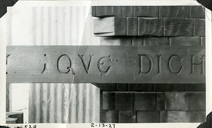 This is an historic black and white photograph from the Scotty's Castle Historic Photograph Collection, Death Valley National Park of the detail of beam before installation in Scotty's Castle Main House Dining Room Carved with QUE DICHA! Set in front of a stack of similar beams. February 13, 1927. Photographed by Mat Roy Thompson.