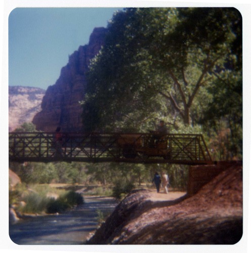 Man driving sweeper equipment across the new Grotto footbridge after its placement on stone abutments. Men working on trail in background.