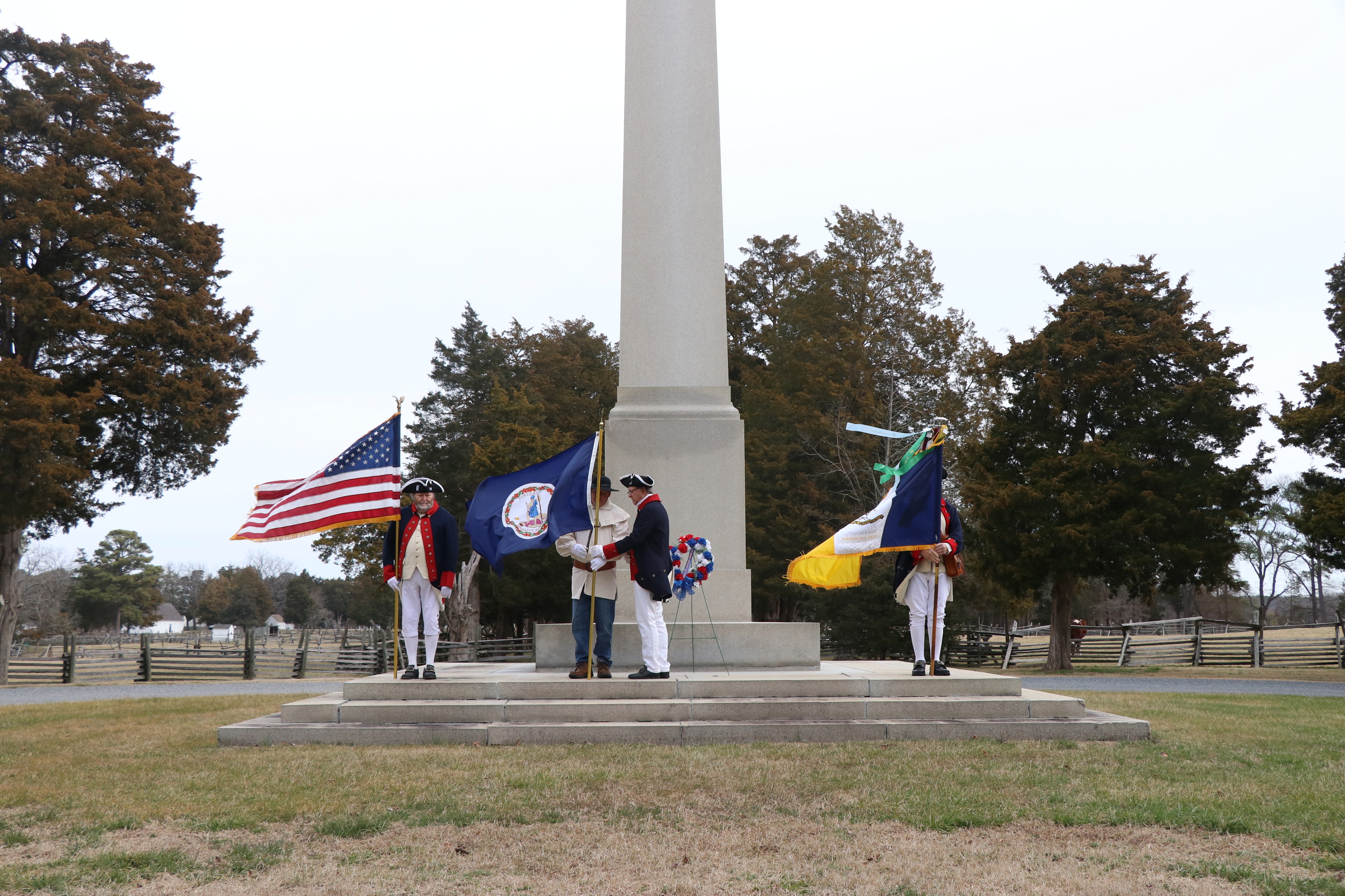 Four men dressed in colonial era clothing, three men holding flags. One reaching out to take one of the flags from the other.