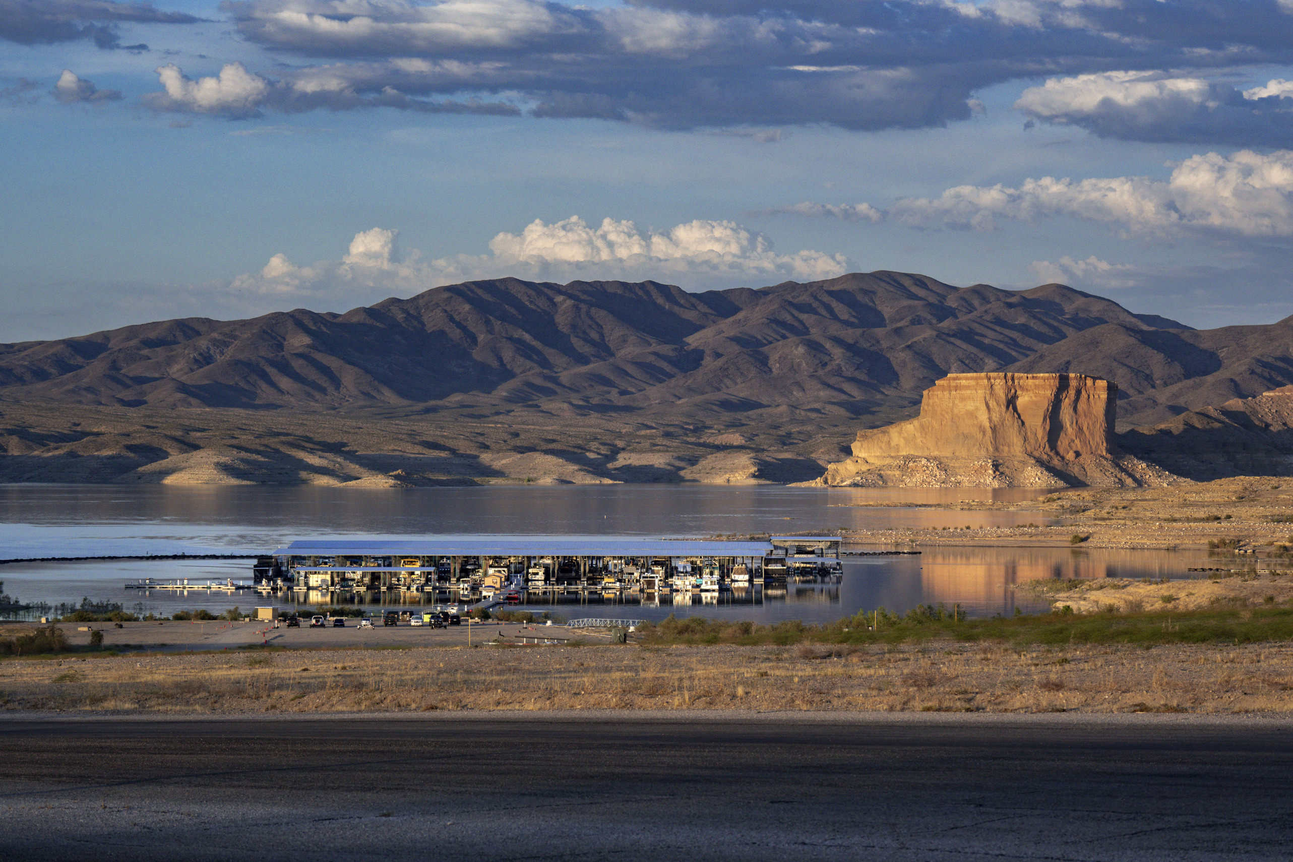 marina viewed from above, surrounded by sandy hills,  flat topped geologic formation behind right, mountains cloudy sky in distance, foliage foreground