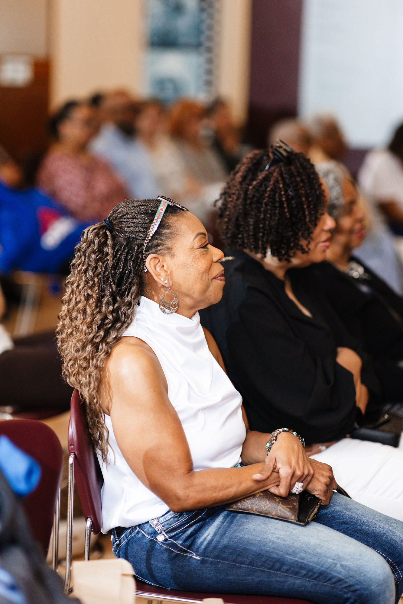 An older african american woman in a white blouse and jeans smiles softly while seated on a maroon plastic chair, surrounded by other african american audience memebers