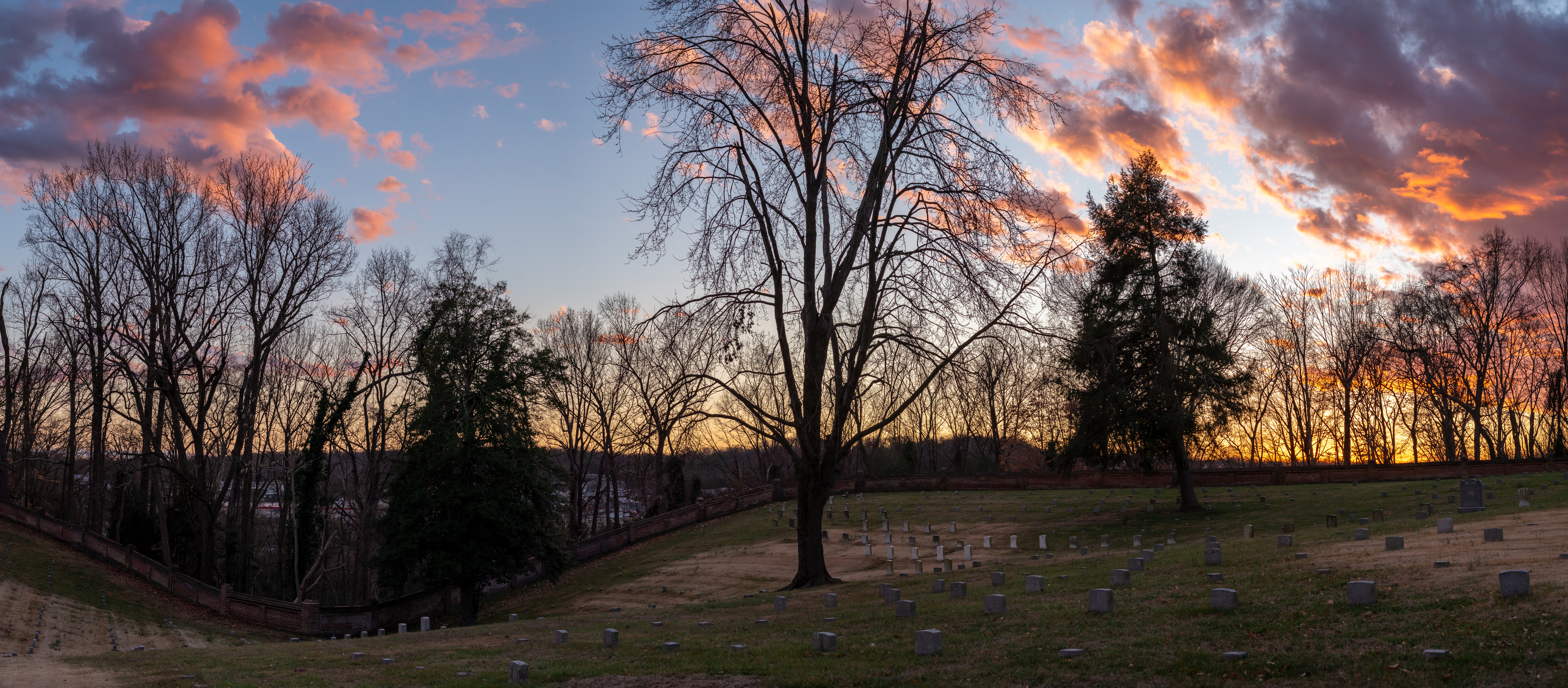 Panorama of a cemetery with sparsely placed trees and a colorful sunset.