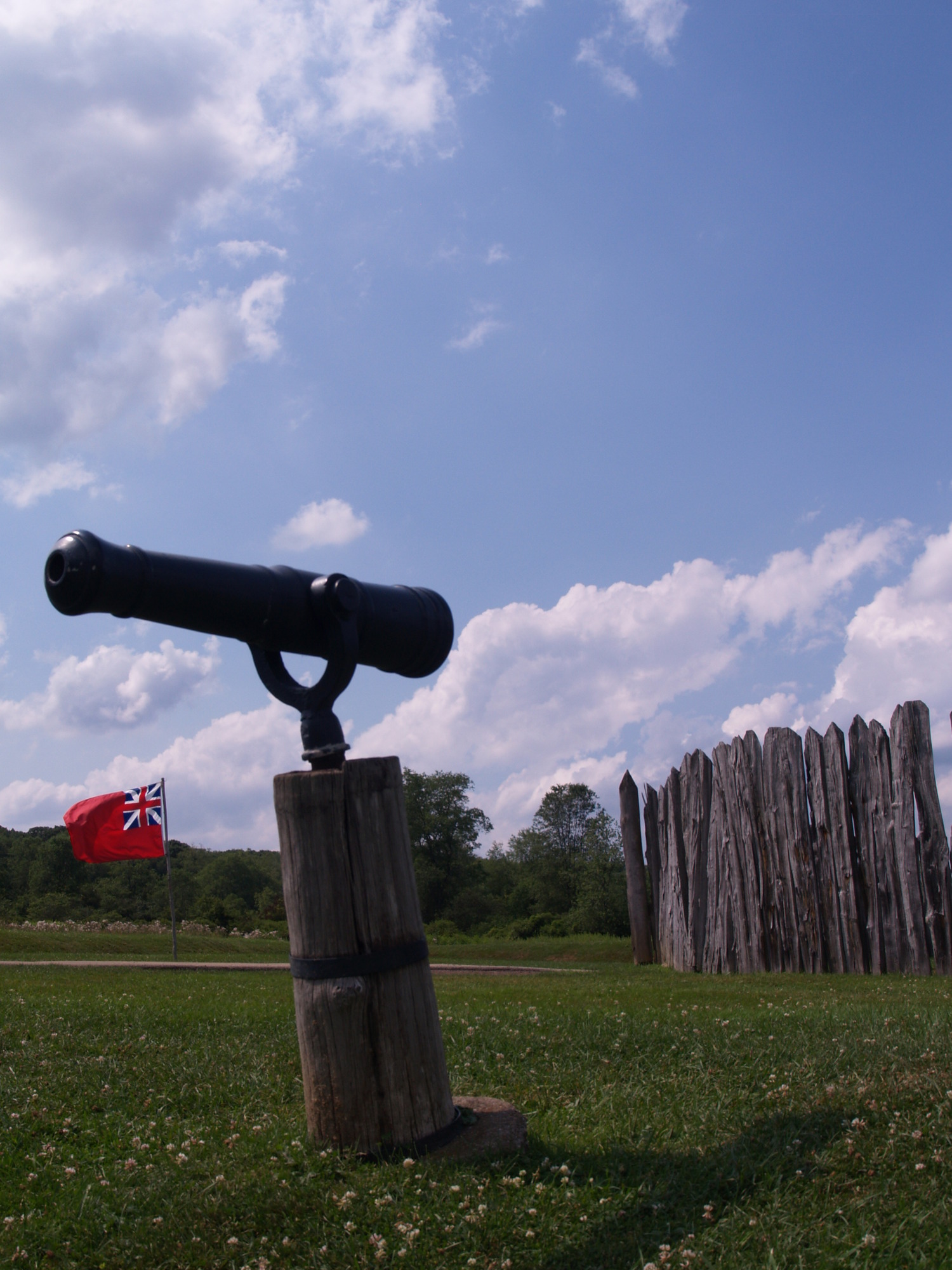 A small piece artillery mounted on a post with the stockade of Fort Necessity and a British navel ensign (flag) waving in the background.