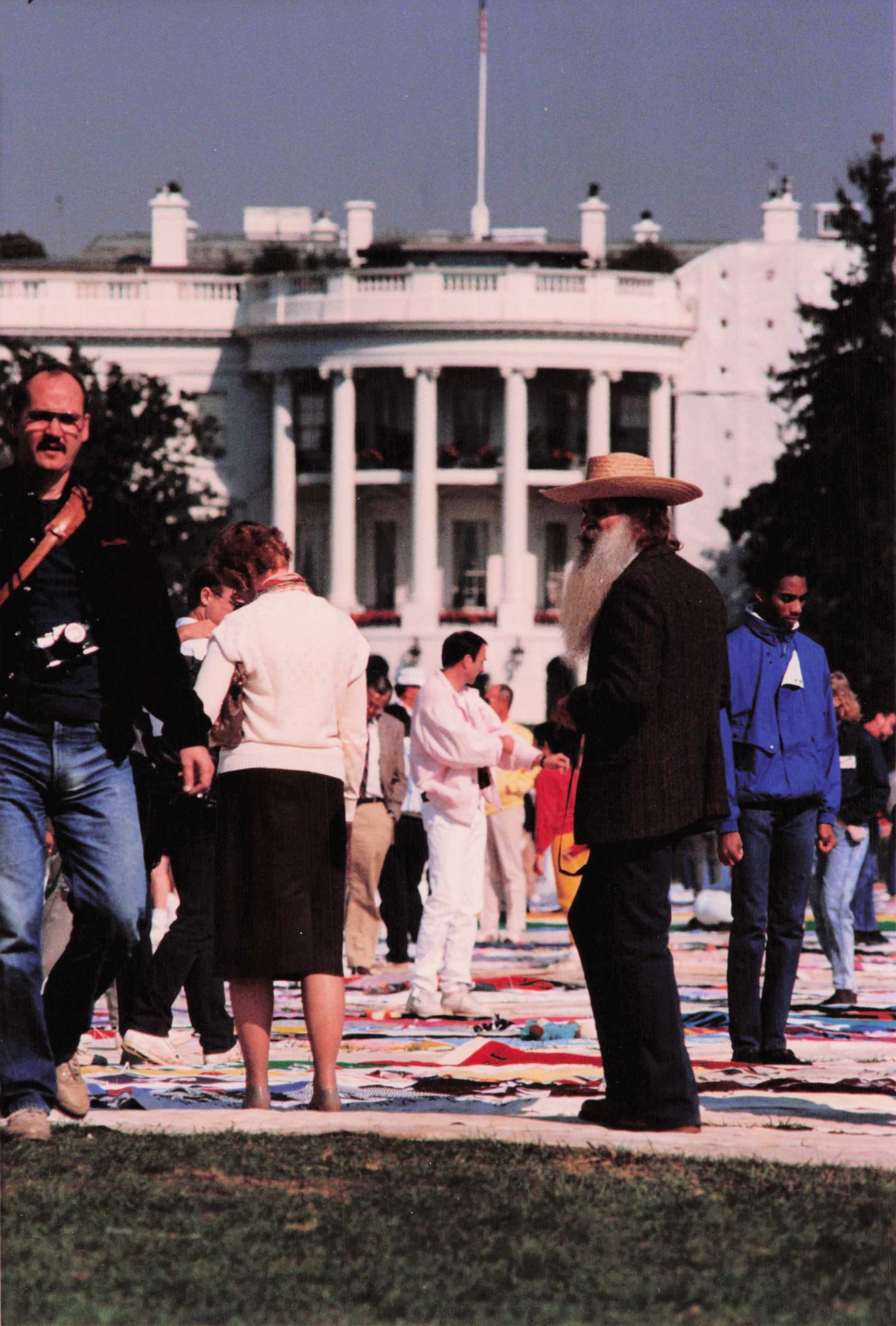 A collection of people stands on multi-colored cloth laid on the grass. They are looking down at the cloth. Trees and the south lawn of the White House are visible in the background