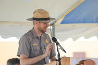 Uniformed ranger smiles as he speaks into the microphone. He stands behind a podium with an NPS arrowhead logo.