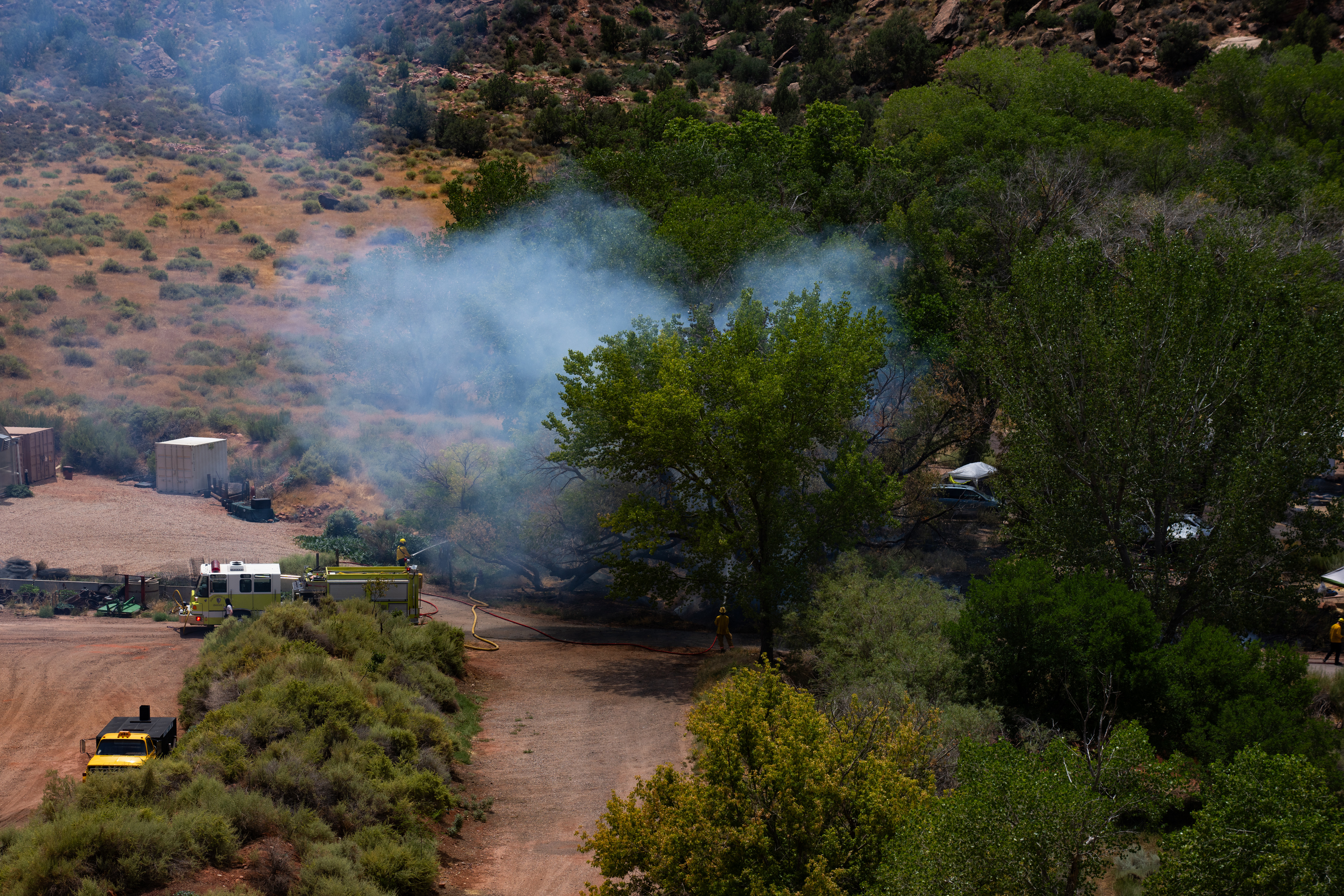 Smoke rises from a wildfire in vegetated area of campground with a fire truck and firefighters working nearby.