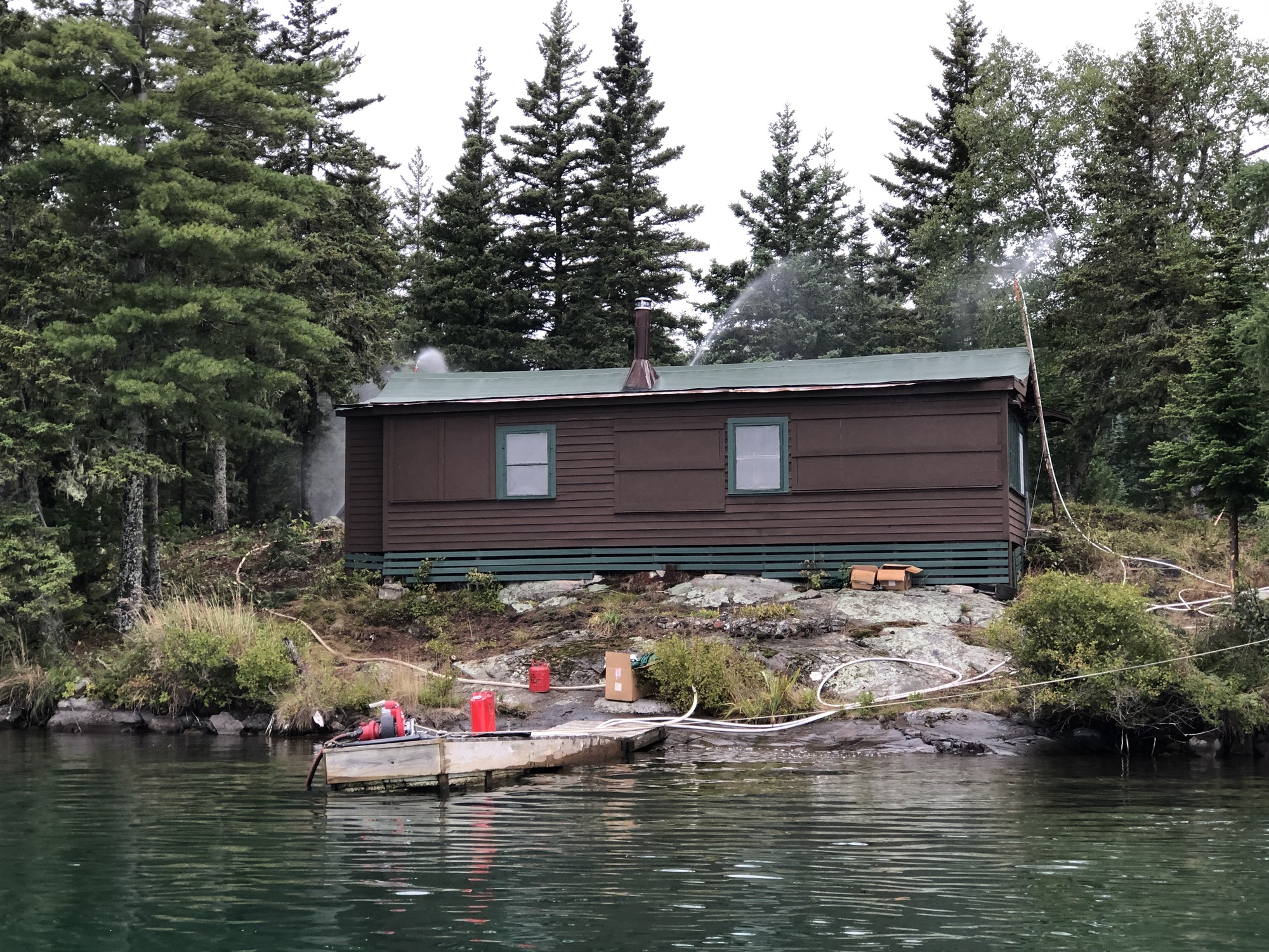 Water view of a small brown and green cabin next to a dock. Sprinklers spray water on and around the building.