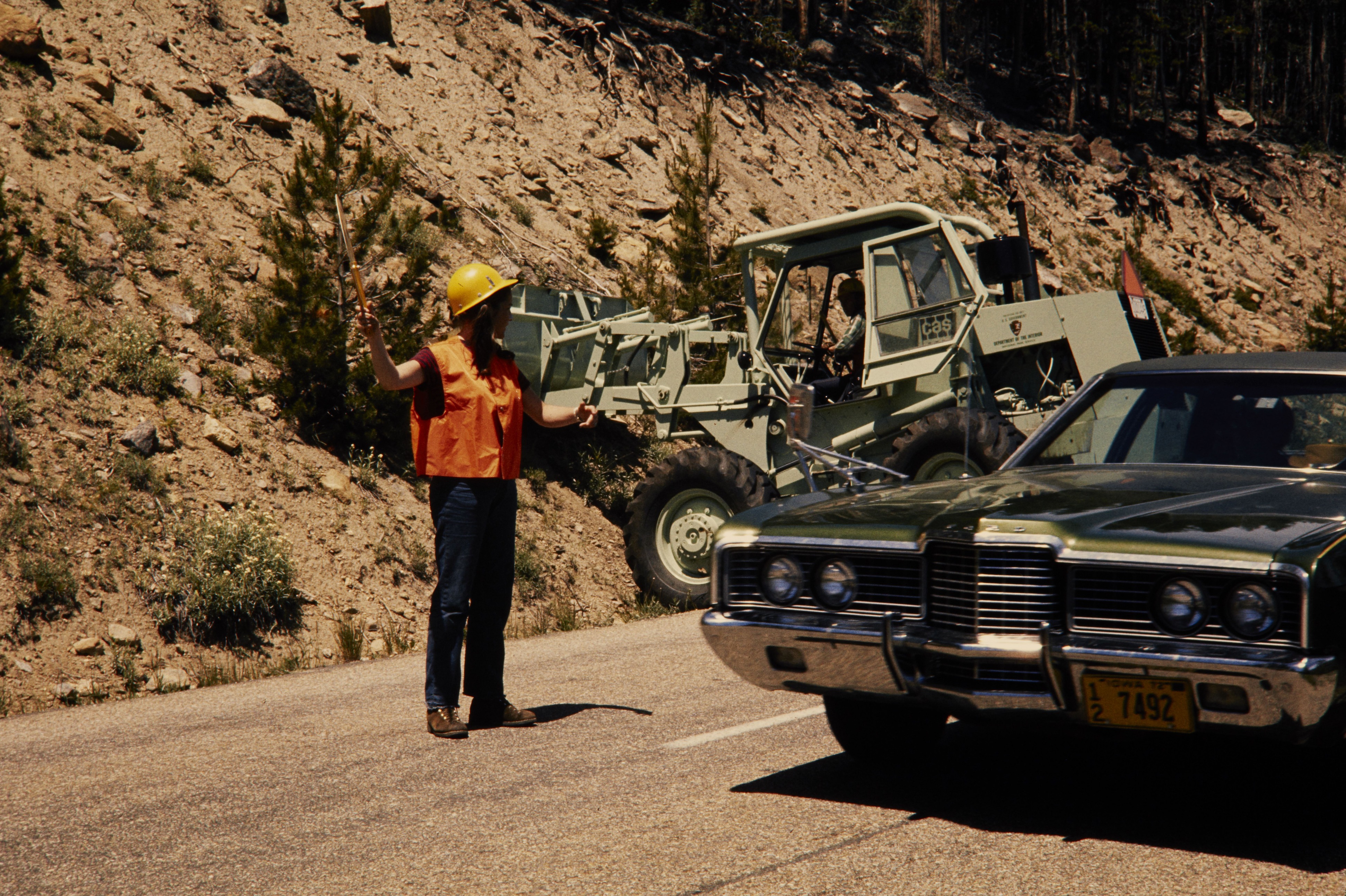 Woman in an orange safety vest and yellow hard hat directs traffic on a road as a backhoe works in the background.