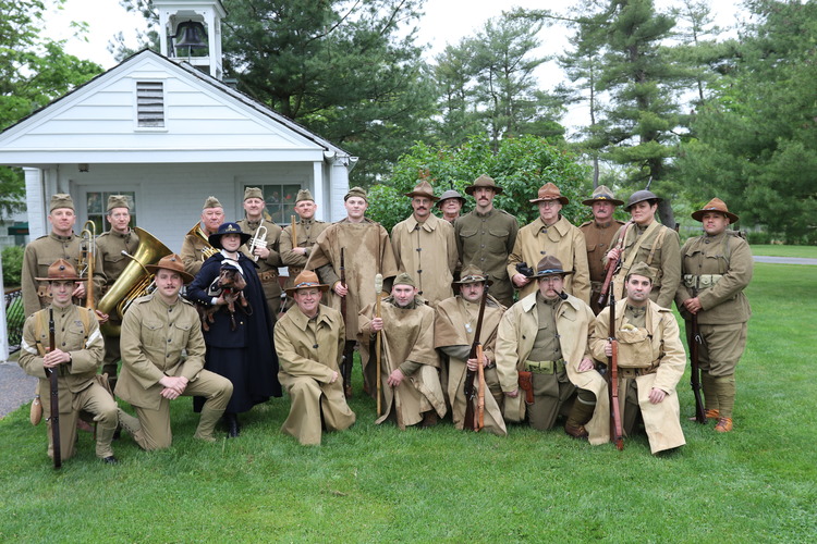 A group of individuals dressed in attire from the WWI era, soldiers and a nurse. A small white building is behind the group. 