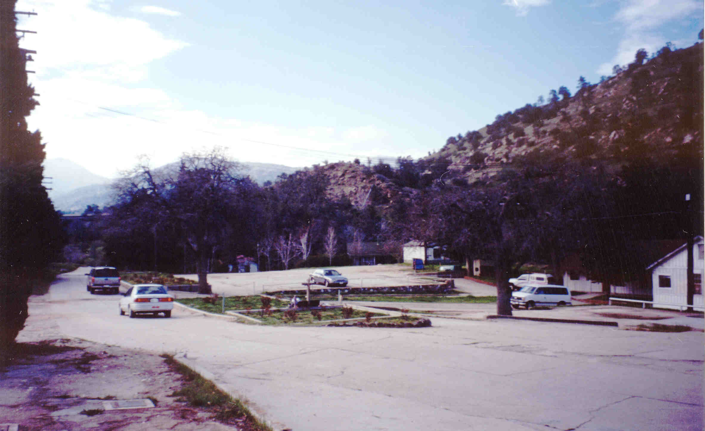 A faded photo shows cars driving near a flat grassy area with a low cross and small bushes.