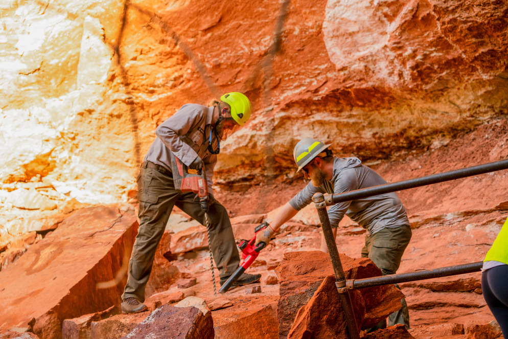 Two maintenance workers in hard hats drill into orange sandstone rock.