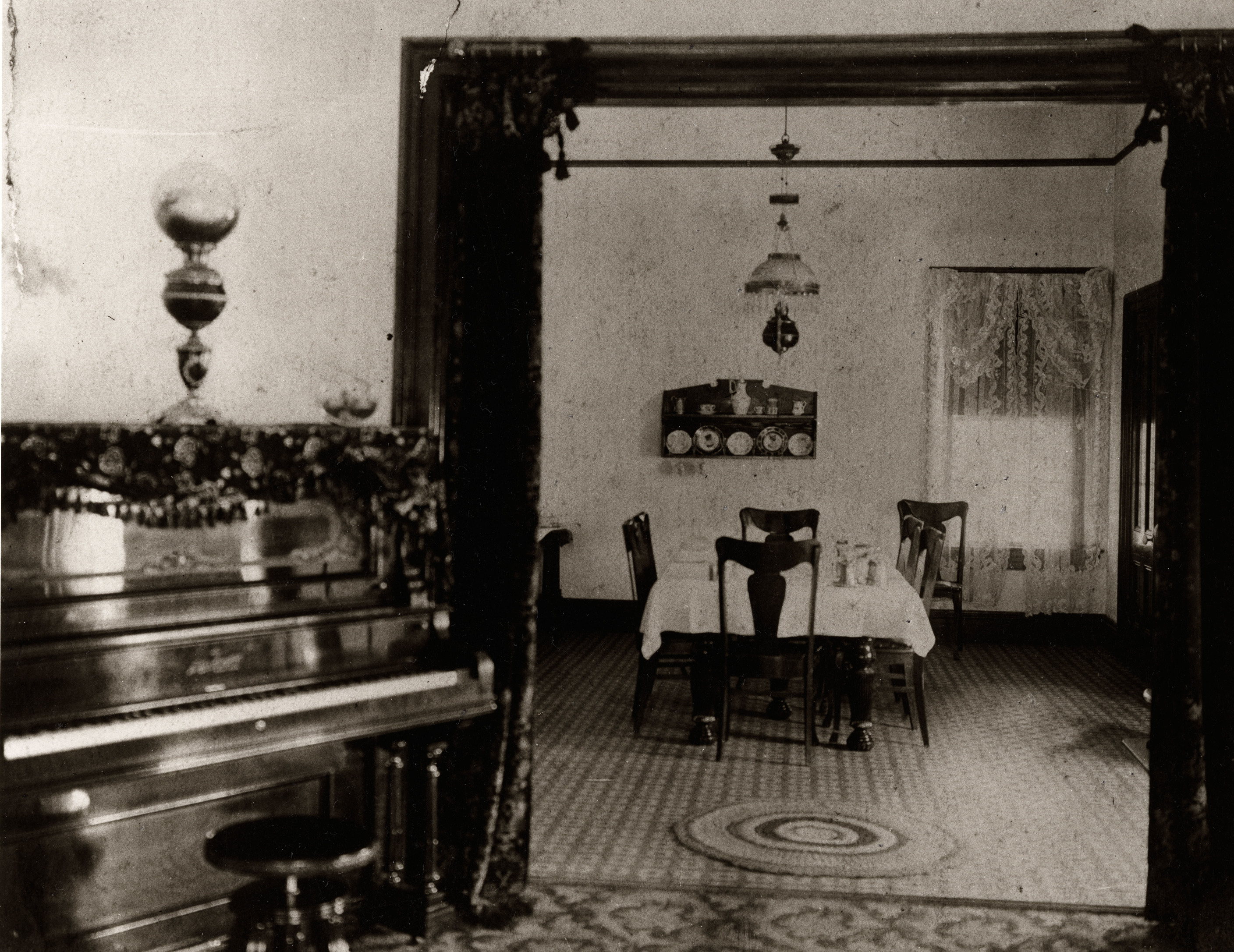 Black and white photograph of rooms inside a house with a dining room table and a piano