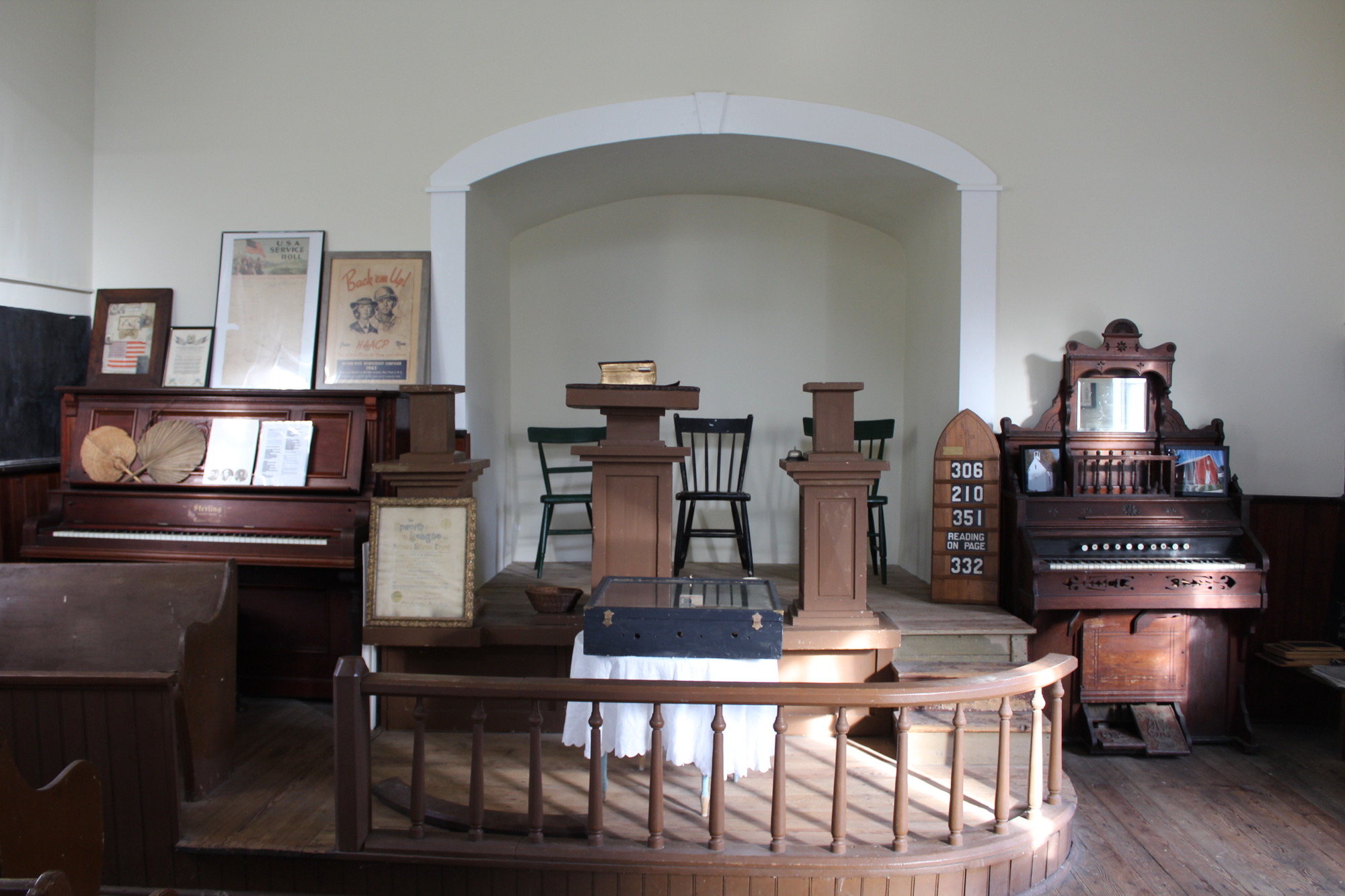 Interior front of a chapel showing plain wooden pulpit flanked by a piano and organ. A side wall is painted with liquid slate to serve as a chalk board.