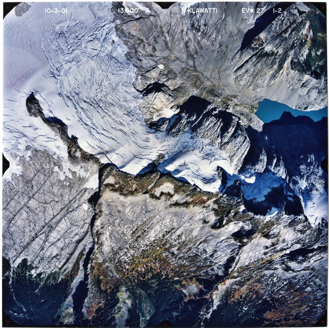 Color aerial photograph of glacier, snowfields, and alpine lake on mountain peak with forested valleys below.