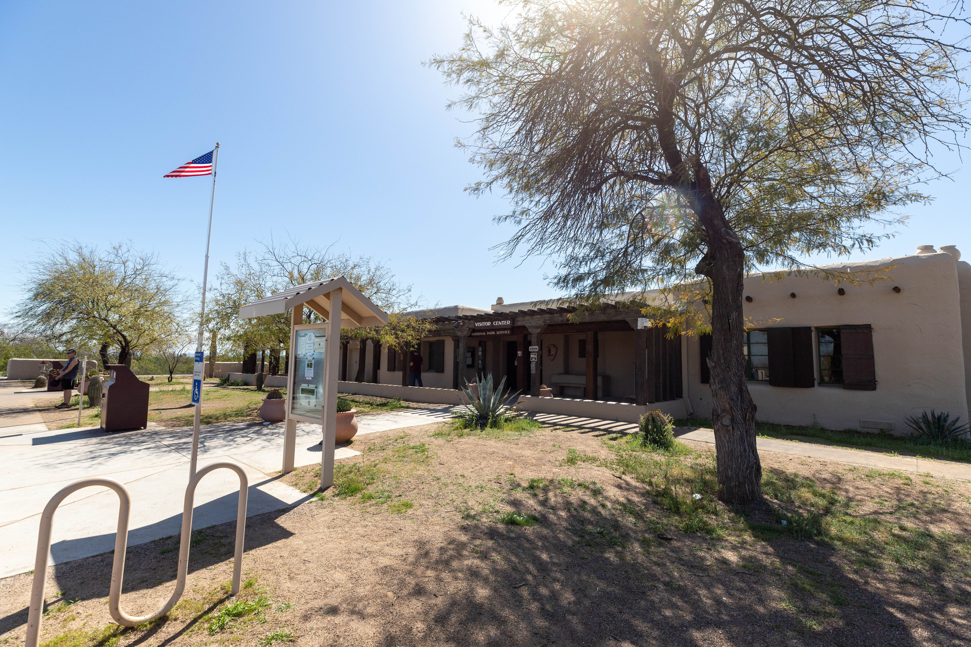 The outdoor space in front of a single story building. There is a flag pole, a tree, a small bike rack, and an information board. 