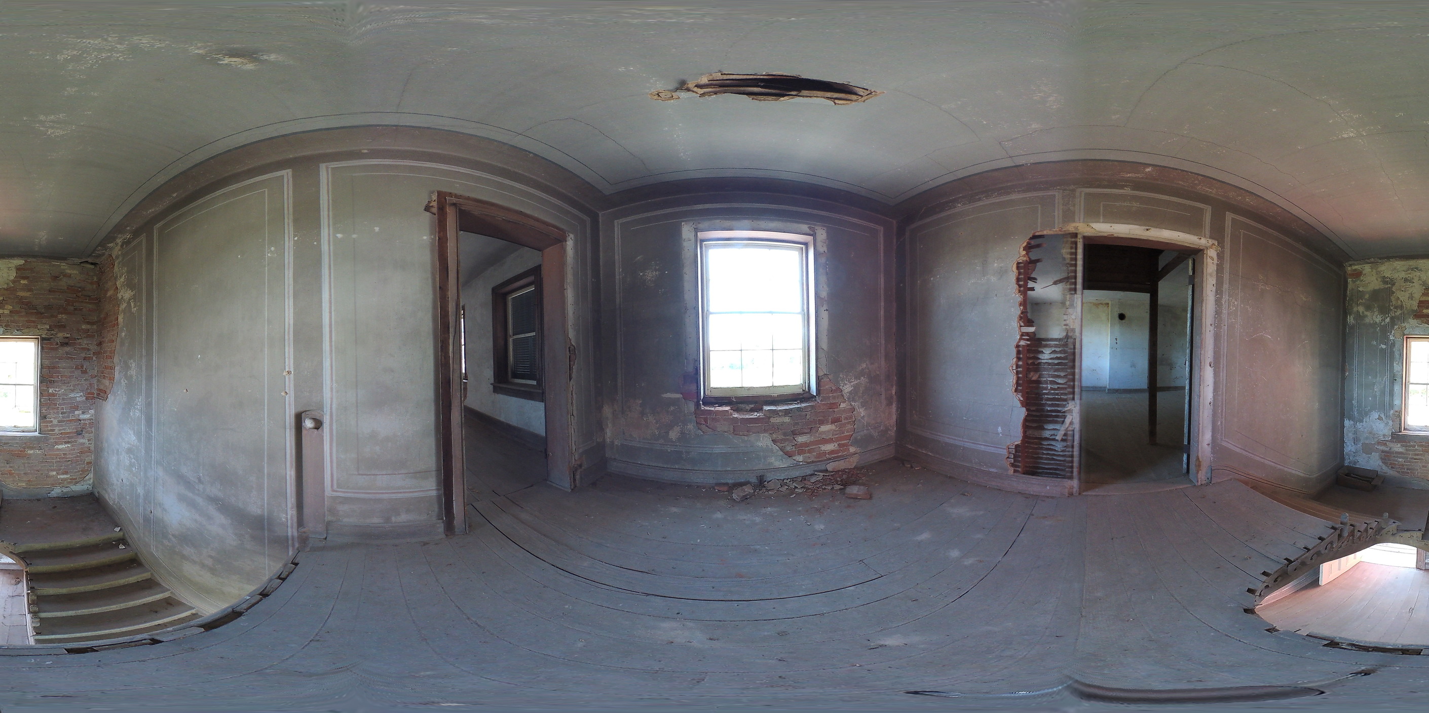 At the top of the stairs, the landing leads to a window. Behind, the staircase leads down to the foyer. Plaster has fallen off the walls, exposing the brick behind. 
