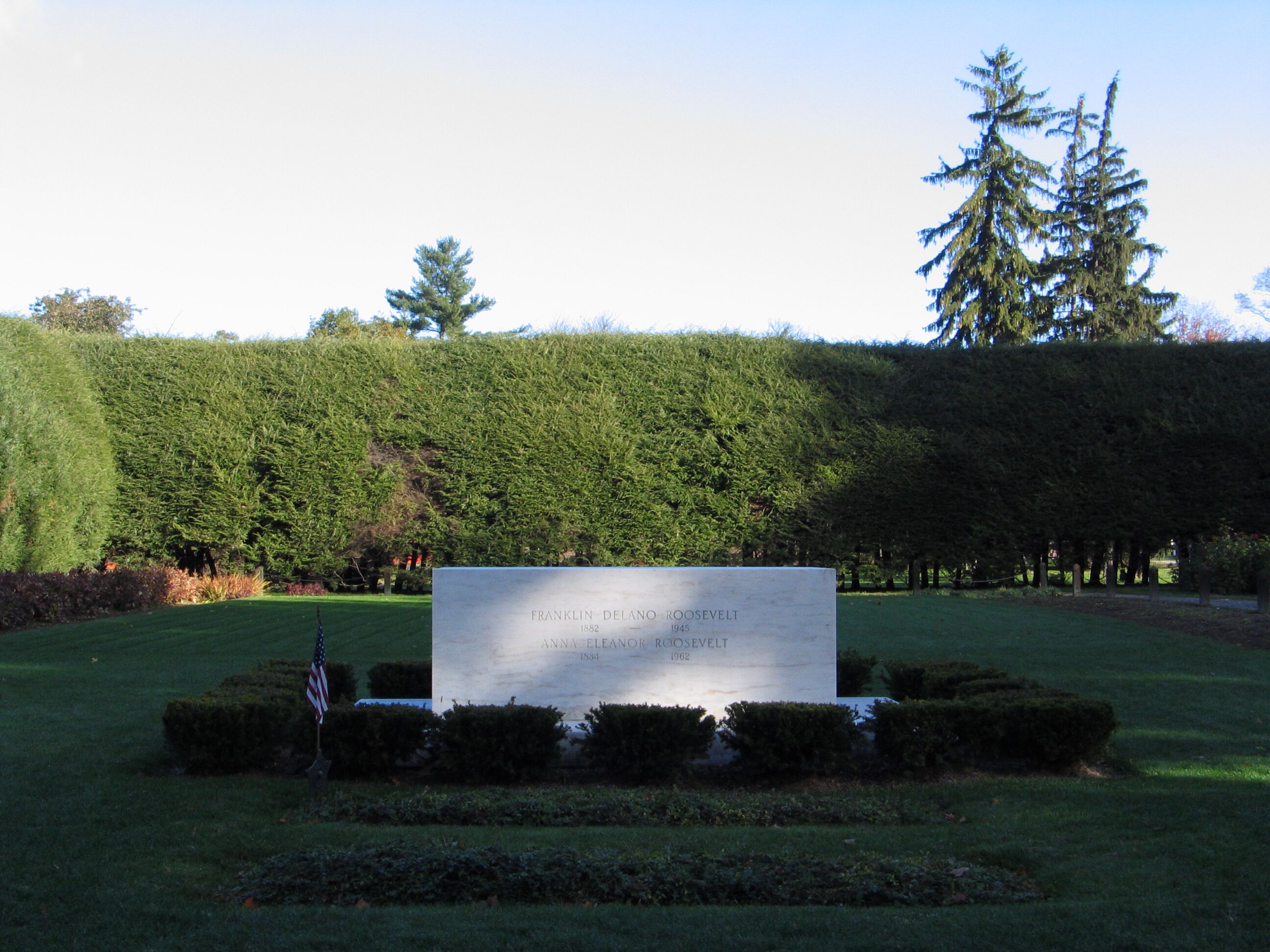 Marble stone in the middle of grass, small American flag in front of and to the left of marble stone, tall hedgerow behind marble stone.