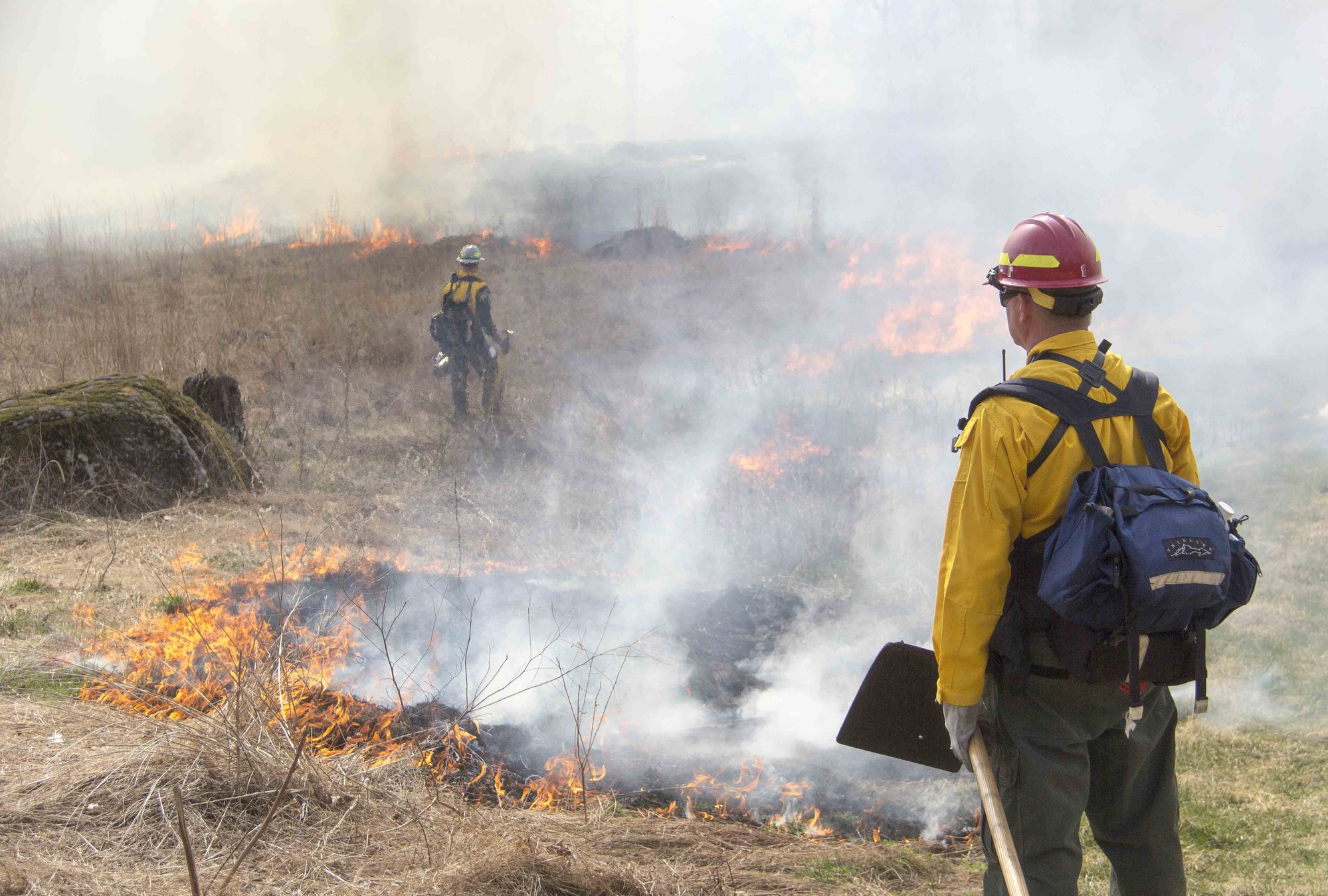Firefighters monitor the prescribed fire as it moves across the fields along the slope of Big Round Top. One firefighter is to the right of the foreground. He is wearing a yellow jacket and hard hat. He is holding a shovel. There is a firefighter in the background. Smoke and fire are present. 