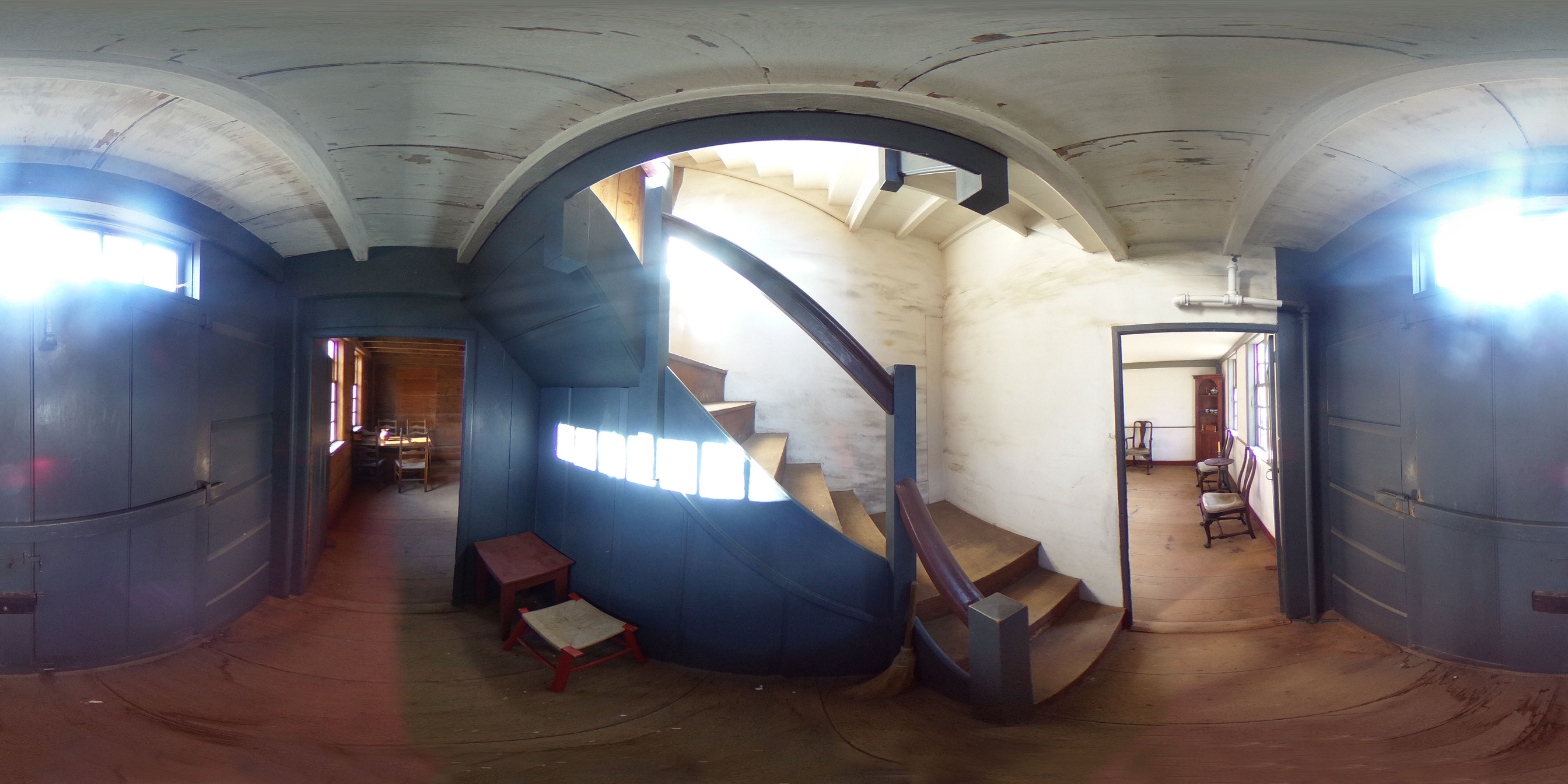360 panoramic image of a colonial house entryway. The main staircase is in front, painted blue/gray. Two rooms are on either side. The walls are white and the floor is unpainted wood. 