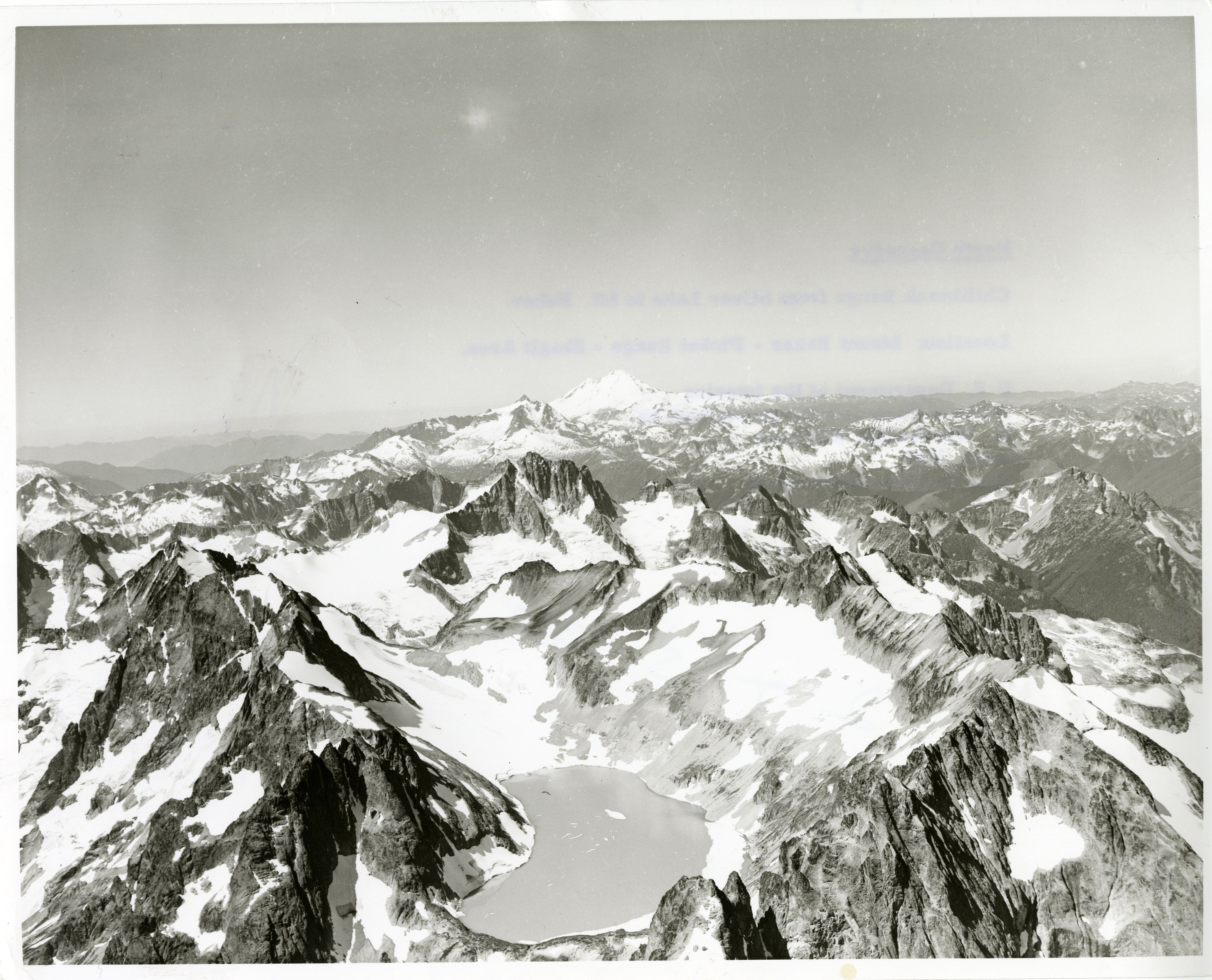 Aerial view of a mountain range. The closest mountain has a lake at the peak. Glaciers, ice and snow can be seen on most mountain tops.