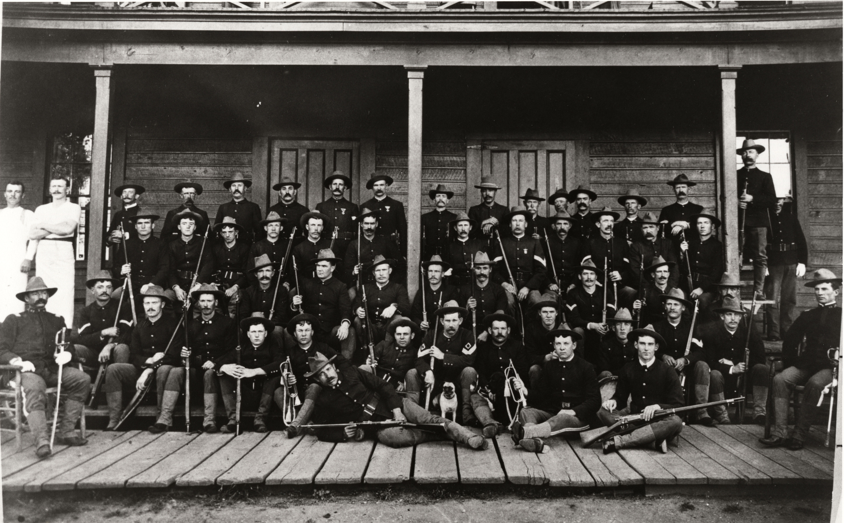 Black and white photograph of rows of soldiers in uniform seated on a porch for a photo