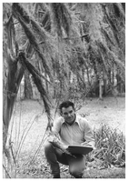 A man wearing long-sleeved shirt kneeling down with a sketchpad beneath Spanish moss and near other tall thin shrubs.