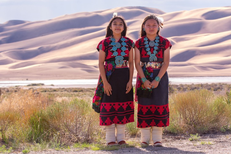 Two Dineh Tah' Navajo Dancers stand in front of the dunes before their presentation at the park Amphitheater. They are wearing colorful traditional black and red dresses and turquoise jewelry. 