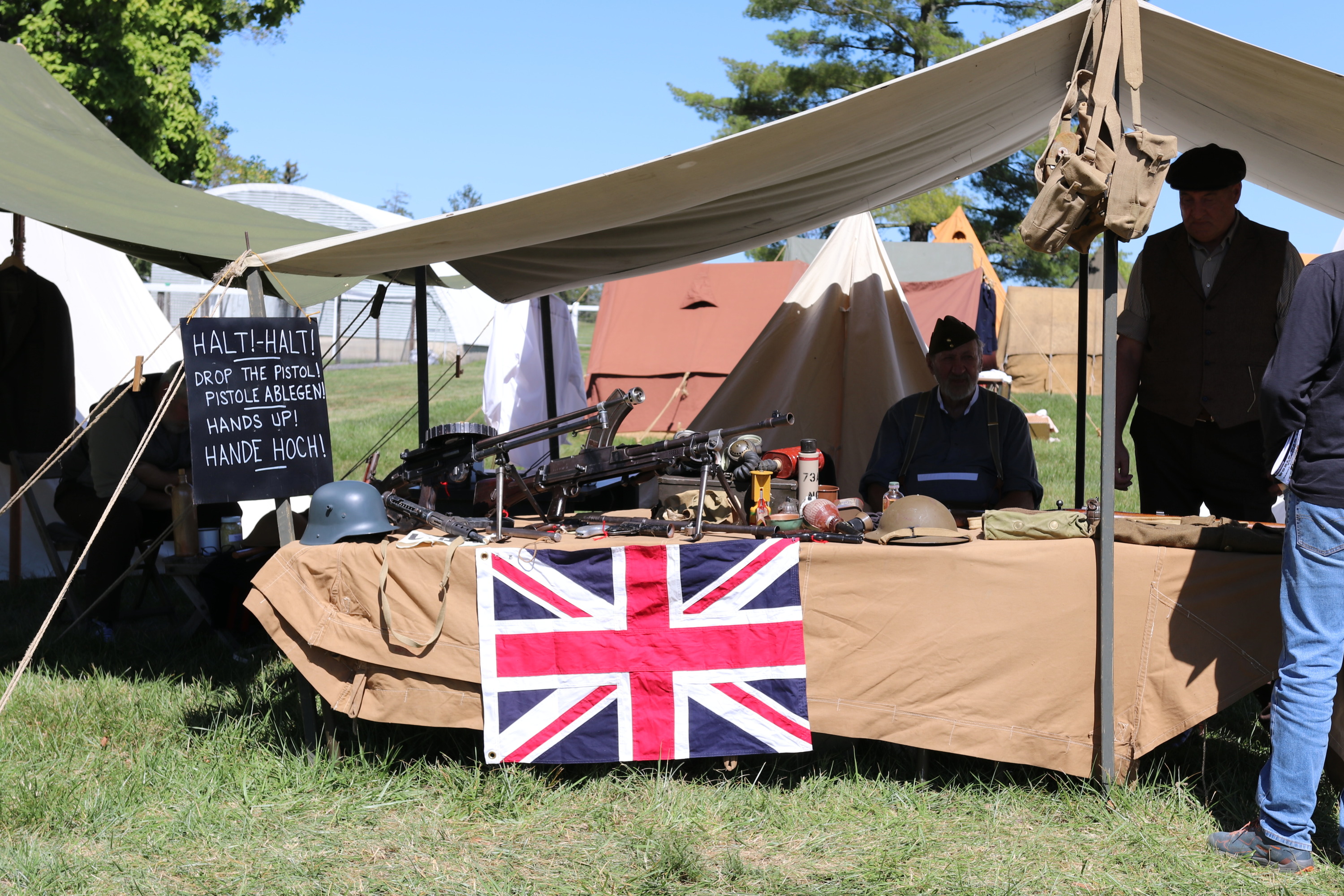 Several living history volunteers sit underneath a khaki tent with a table covered with historic items and a British flag. 