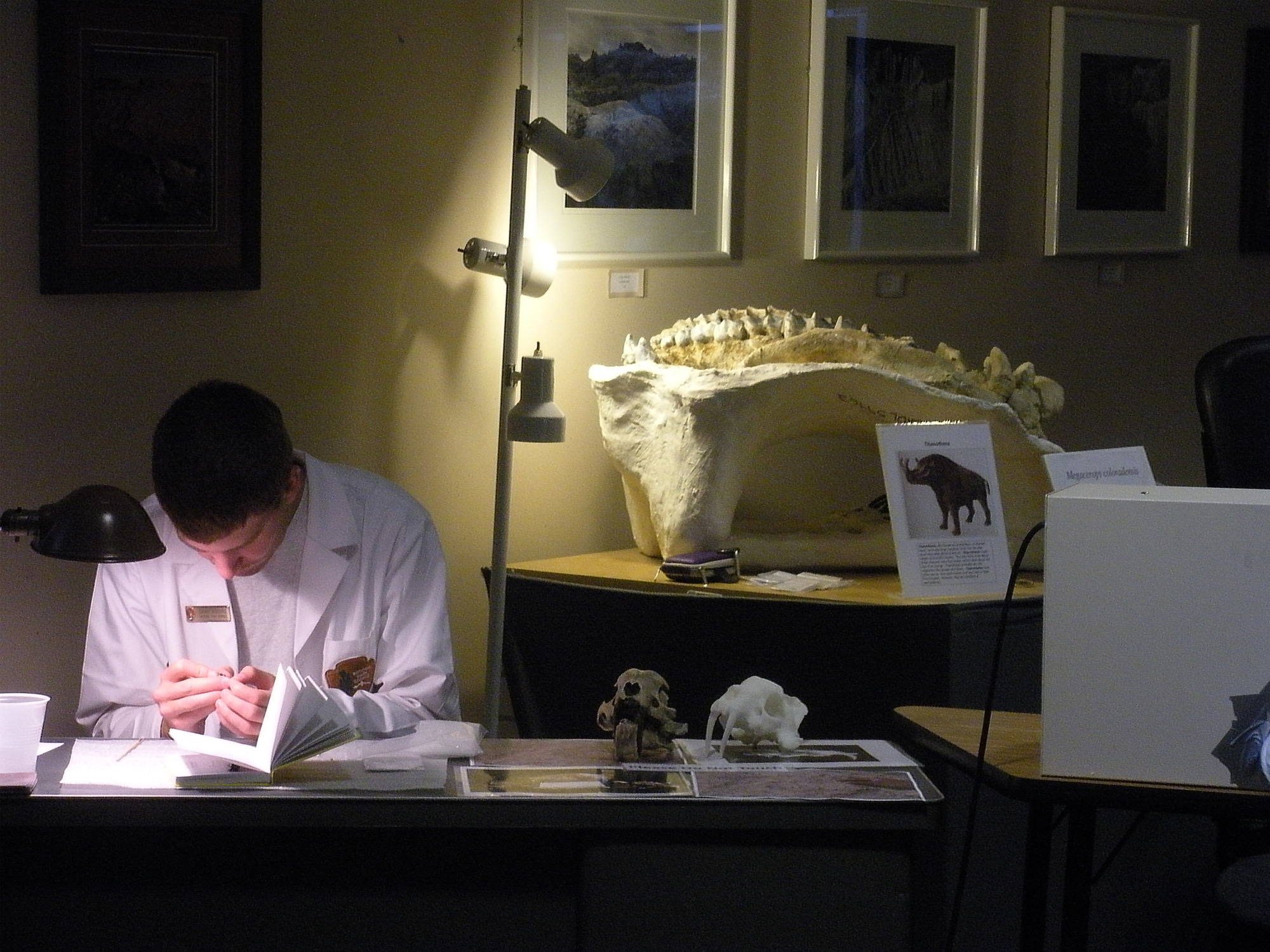 a young man in a white lab coat sits at a desk in a dark room. 