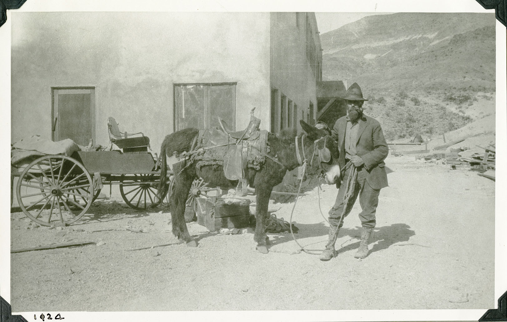 This is an historic black and white photograph from the Scotty's Castle Historic Photograph Collection, Death Valley National Park of a man holding onto a saddled burro in front of a large structure and wagon. Man is dressed in high topped hat, coat, shirt, and jeans tucked into high-top, laced boots. Burrow had bridle with blinders, western saddle with breast collar and rear britchen. Buckboard style wagon with wooden crates and pack behind burro. Simple, large, two-story stucco building with many windows. Number in black ink in lower left corner.