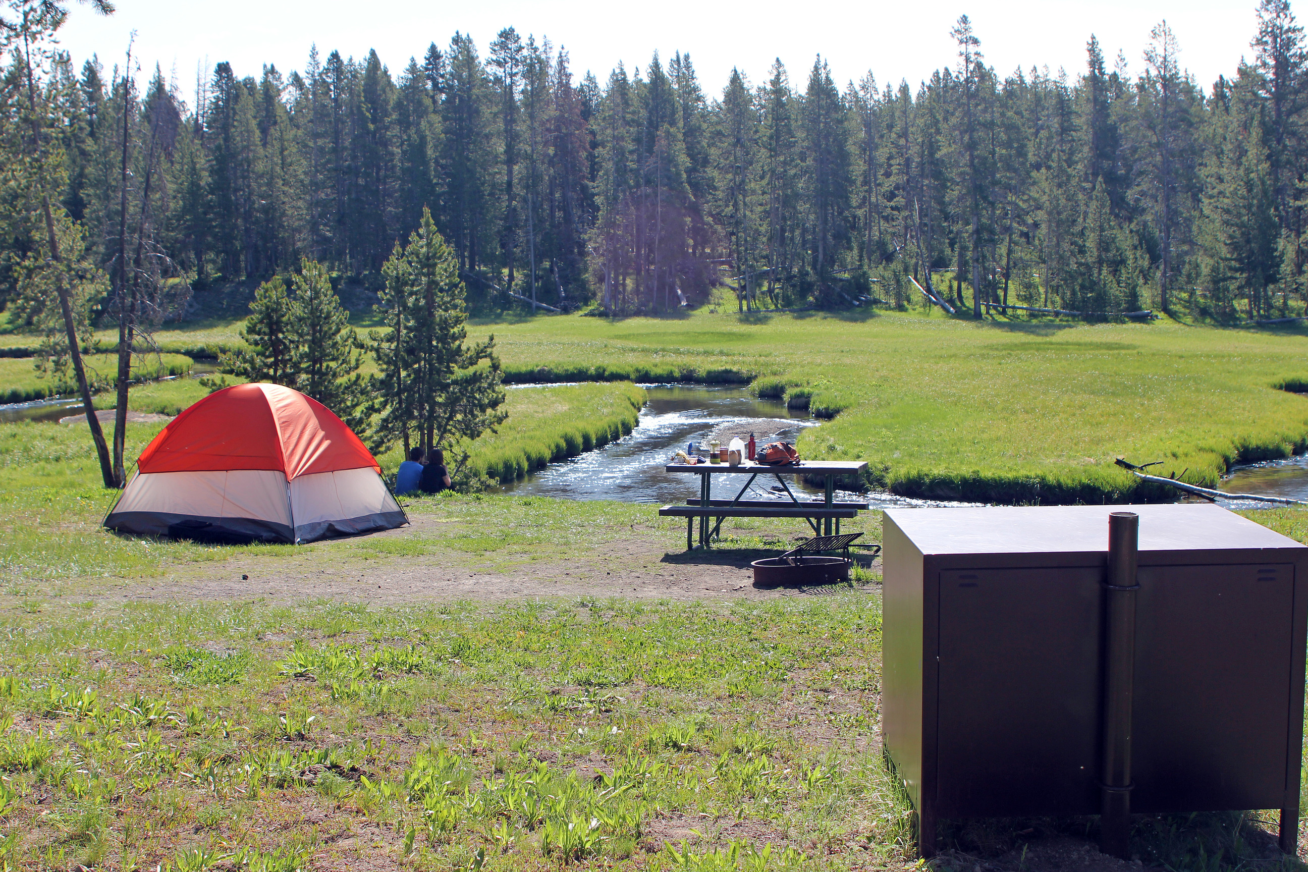 An orange tent is set up next to a picnic table which is next to a river.