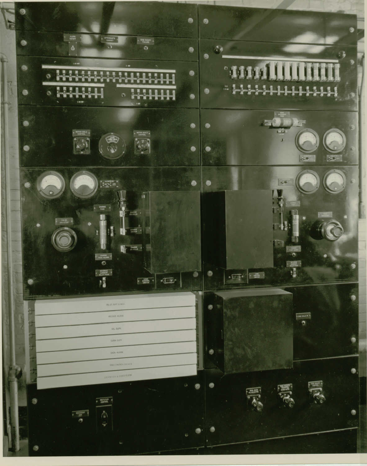 Black and white photo of a large electrical panel. Several switches, knobs, and gauges with labels cover the panel.
