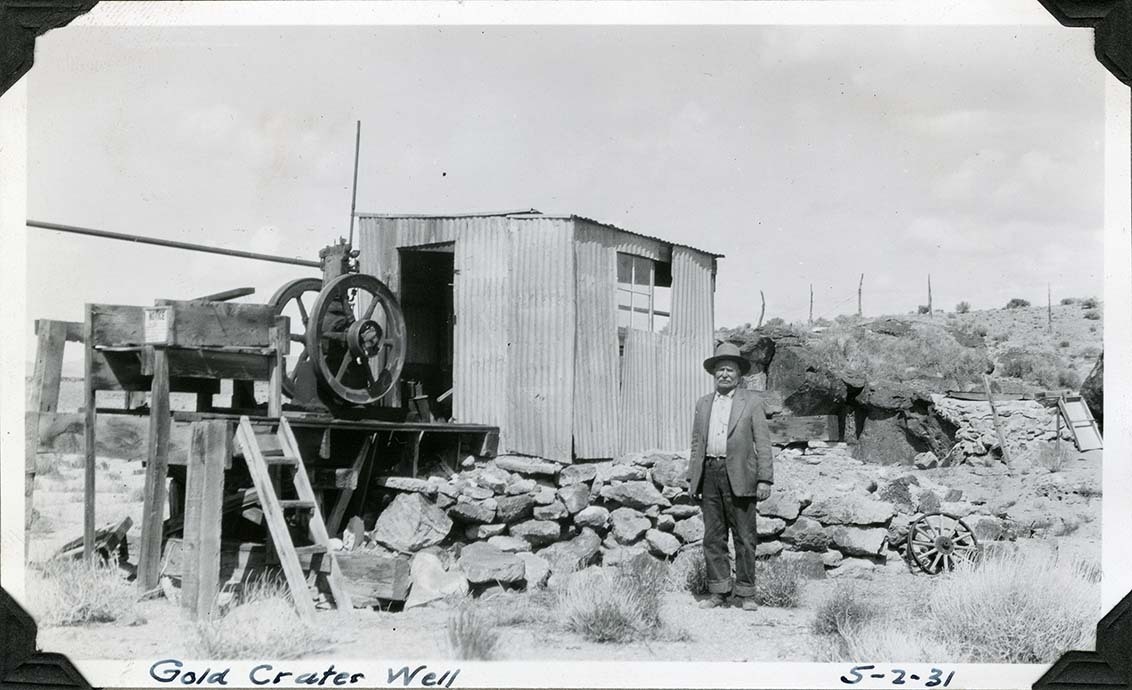 This is an historic black and white photograph from the Scotty's Castle Historic Photograph Collection, Death Valley National Park of man posing for camera in front of metal shack on rock foundation with pump equipment on deck in front. Shack is made of corrugated tin. Wood platform with ladder from ground holds equipment. Metal, two-wheel equipment, probably pump, on platform. Man dressed in hat, simple shirt, jeans with cuff rolled up.