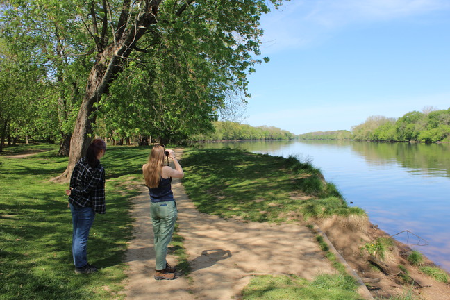 Two people stand on a riverbank looking through binoculars.