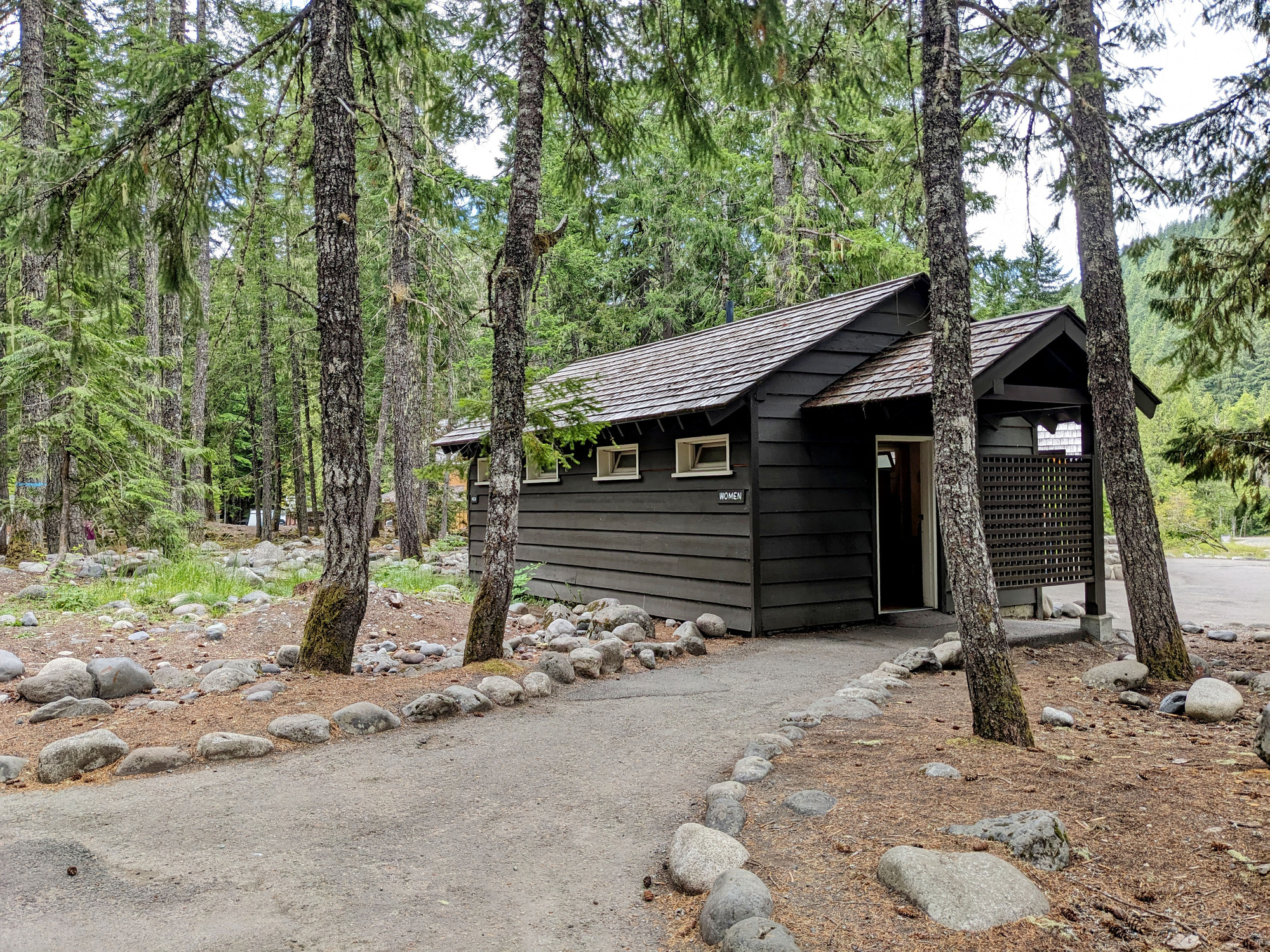 A stone-lined sidewalk leads to a dark brown restroom building in the forest.