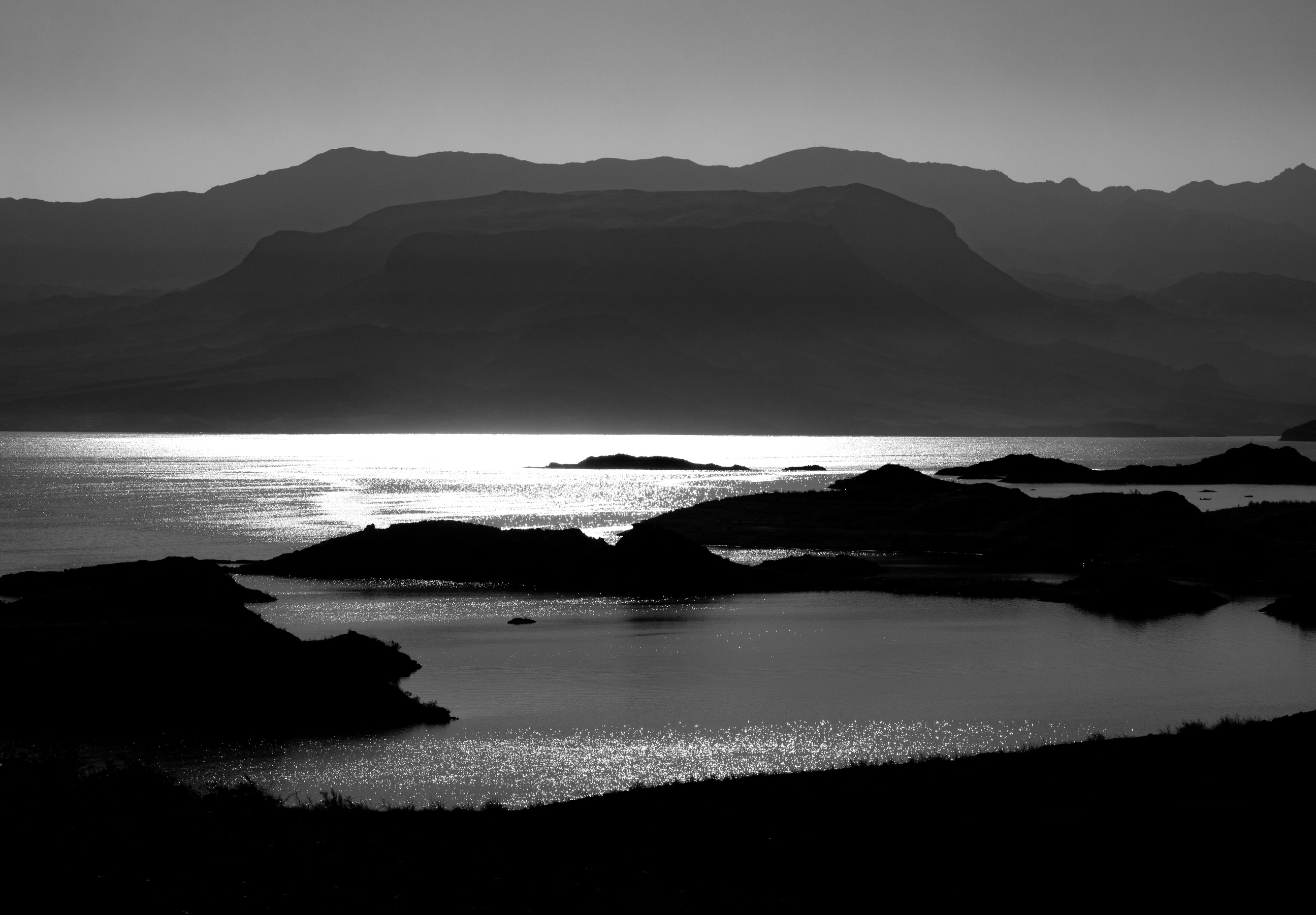 monochrome lake with bright reflection in center surrounded by hazy mountains and small islands