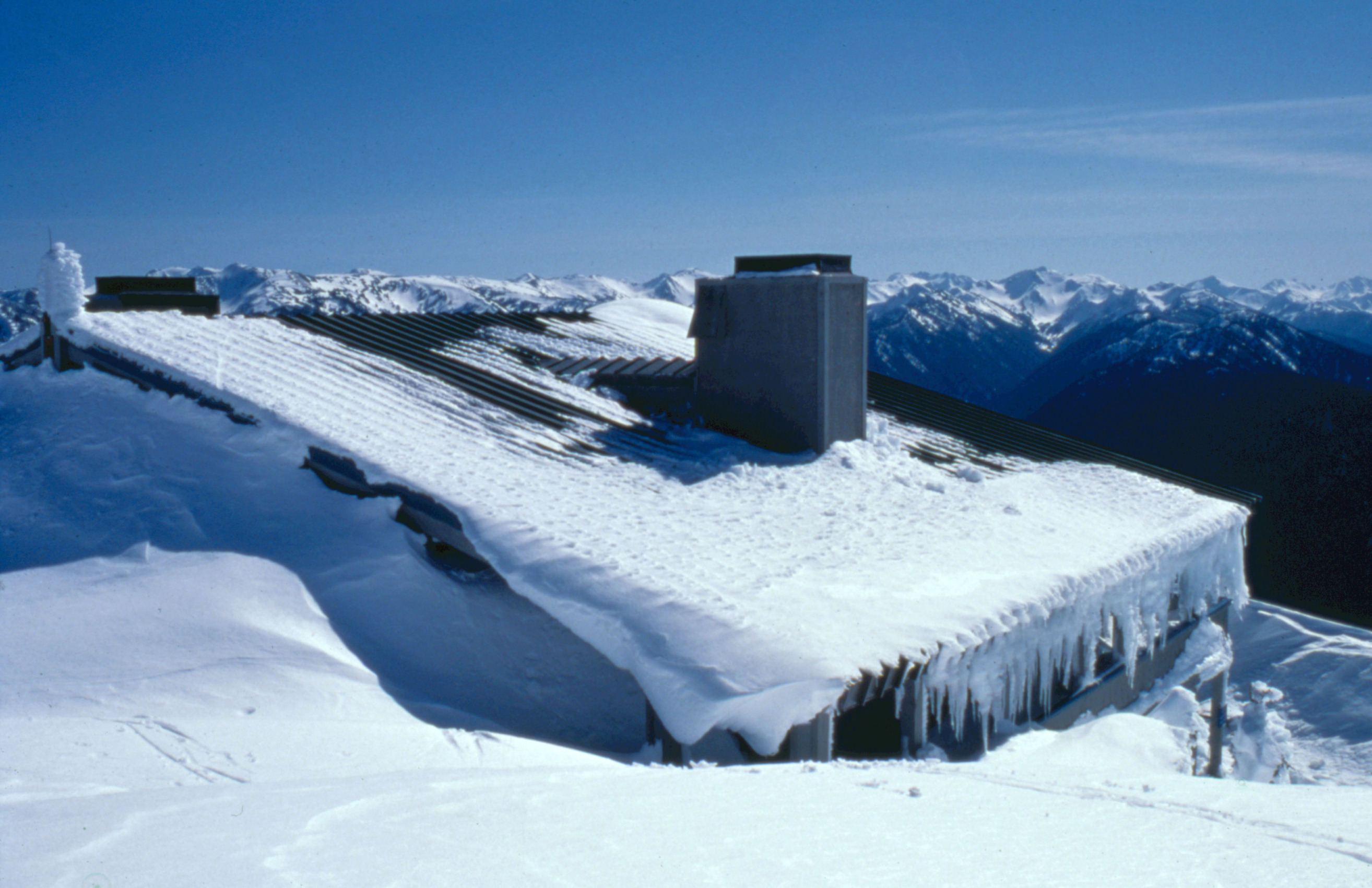 A building nearly hidden in drifted snow, with its roof visible and icicles. Snowy mountains in the distance.