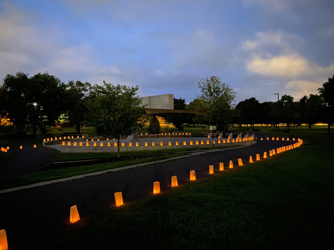 Dozens of lighted luminaria bags surround a large brass bell in a city park.
