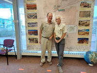 Uniformed man and woman in front of a large wall map and windows inside.
