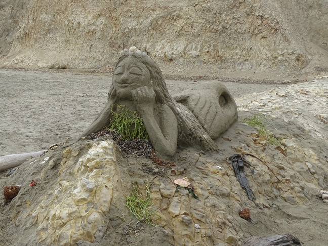 A sand sculpture of a mermaid lying on her belly on top of a rock outcrop with her elbows on the rock and head propped up in her hands.