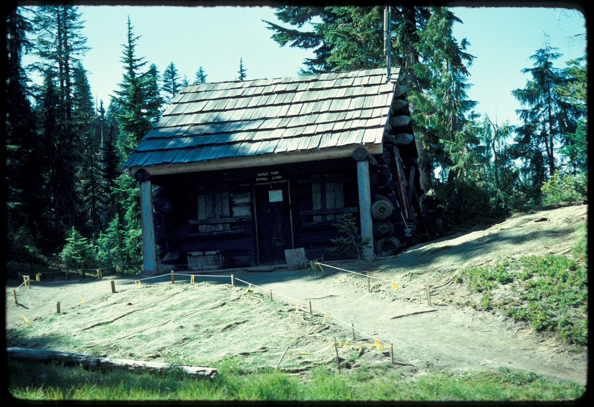 A small log cabin with a covered porch. Burlap netting covers the vegetation in front of the cabin on either side of the path to the entrance. 