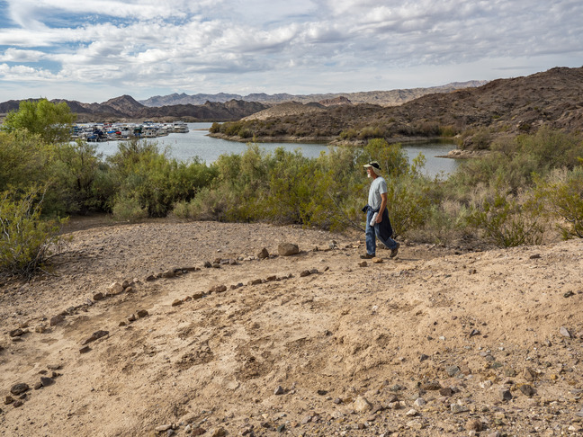 rock lined sandy trail, man walking on trail at right, foliage lake marina and mountains in distance
